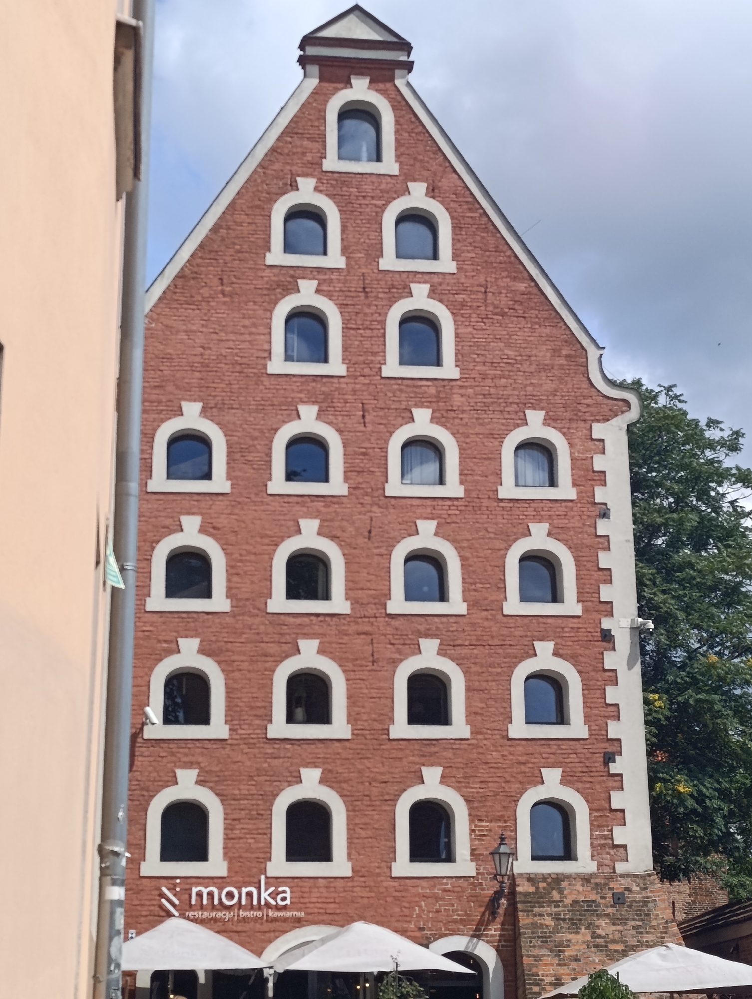 The image captures the facade of a brick building with a gabled roof in Toruń, Poland. The building features multiple rows of arched windows with decorative white surrounds. A sign with the text "monka restauracja bistro kawiarnia" is visible on the lower part of the building, indicating it is a restaurant, bistro, and cafe. White awnings or umbrellas are set up in front of the establishment, suggesting an outdoor seating area. To the left of the brick building, a portion of a lighter-colored building is visible. The sky above is partly cloudy. The overall scene depicts a commercial establishment in an urban setting.