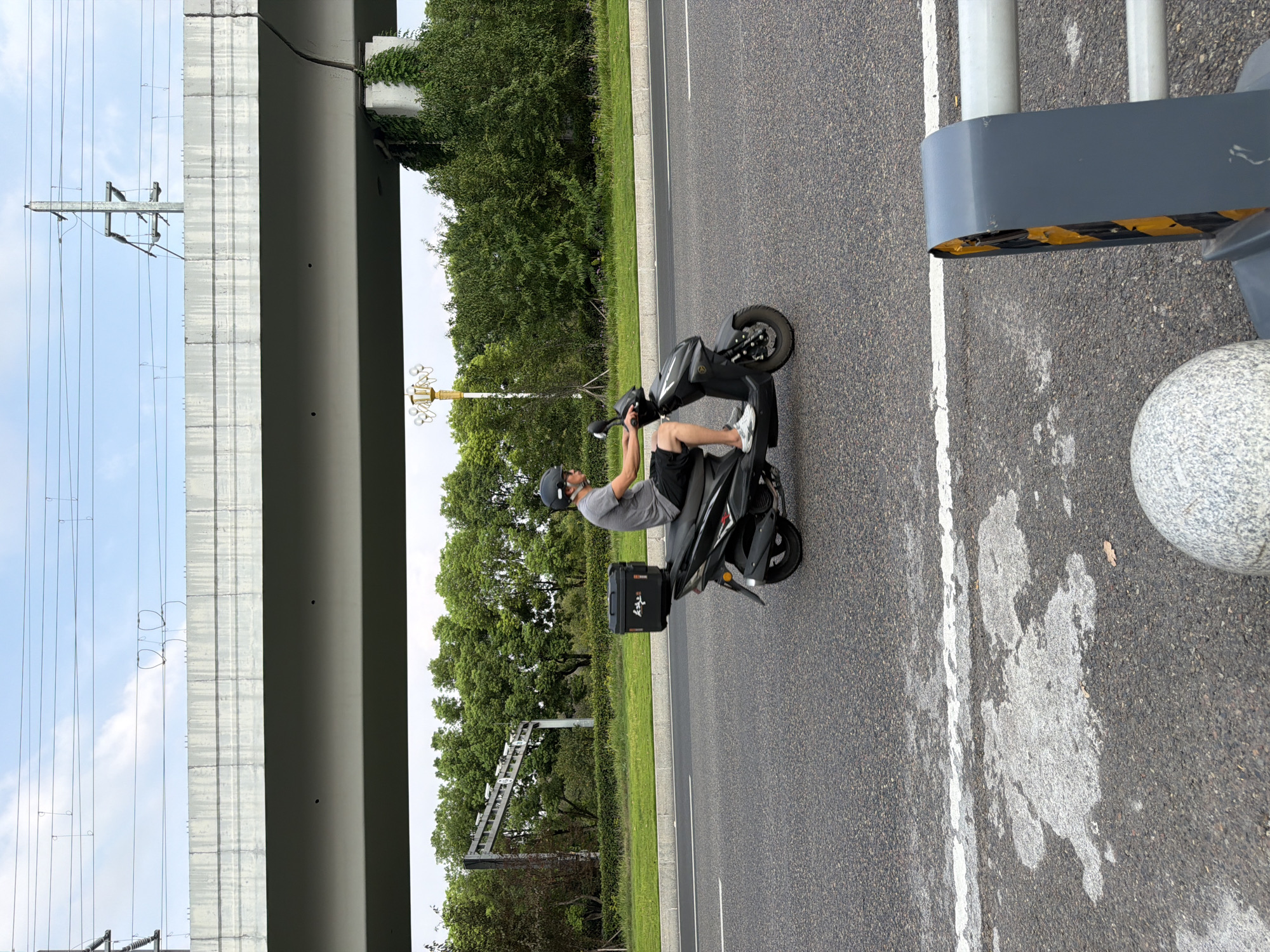 A man wearing a helmet, grey t-shirt, and shorts rides a black scooter on a paved road. The scooter has a black storage box mounted on the back. The road is bordered by a green grassy area and trees. Above the trees is a concrete overpass with power lines running across it. The sky is blue with scattered clouds. The image appears to be taken on a sunny day. The setting is likely a city in Jiangjin, China, given the presence of overpasses and electrical infrastructure common in urban areas.