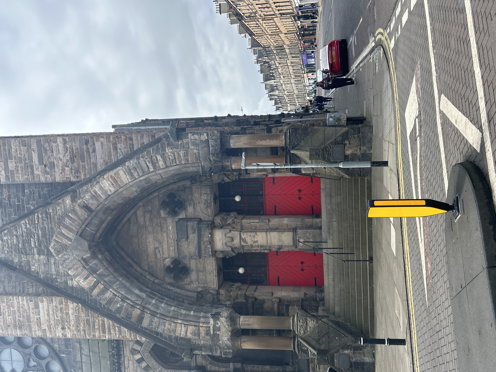 A medium shot captures the imposing stone facade of a church with two prominent red doors leading up a set of concrete stairs. Flanking the doors are substantial stone pillars. To the right of the church, a street stretches into the distance, lined with a row of terraced buildings featuring multi-story facades and numerous windows. A red car is parked on the right side of the street. The pavement in the foreground features a yellow bollard. Several pedestrians are walking along the pavement. The sky is overcast. The image is taken from a street-level perspective.