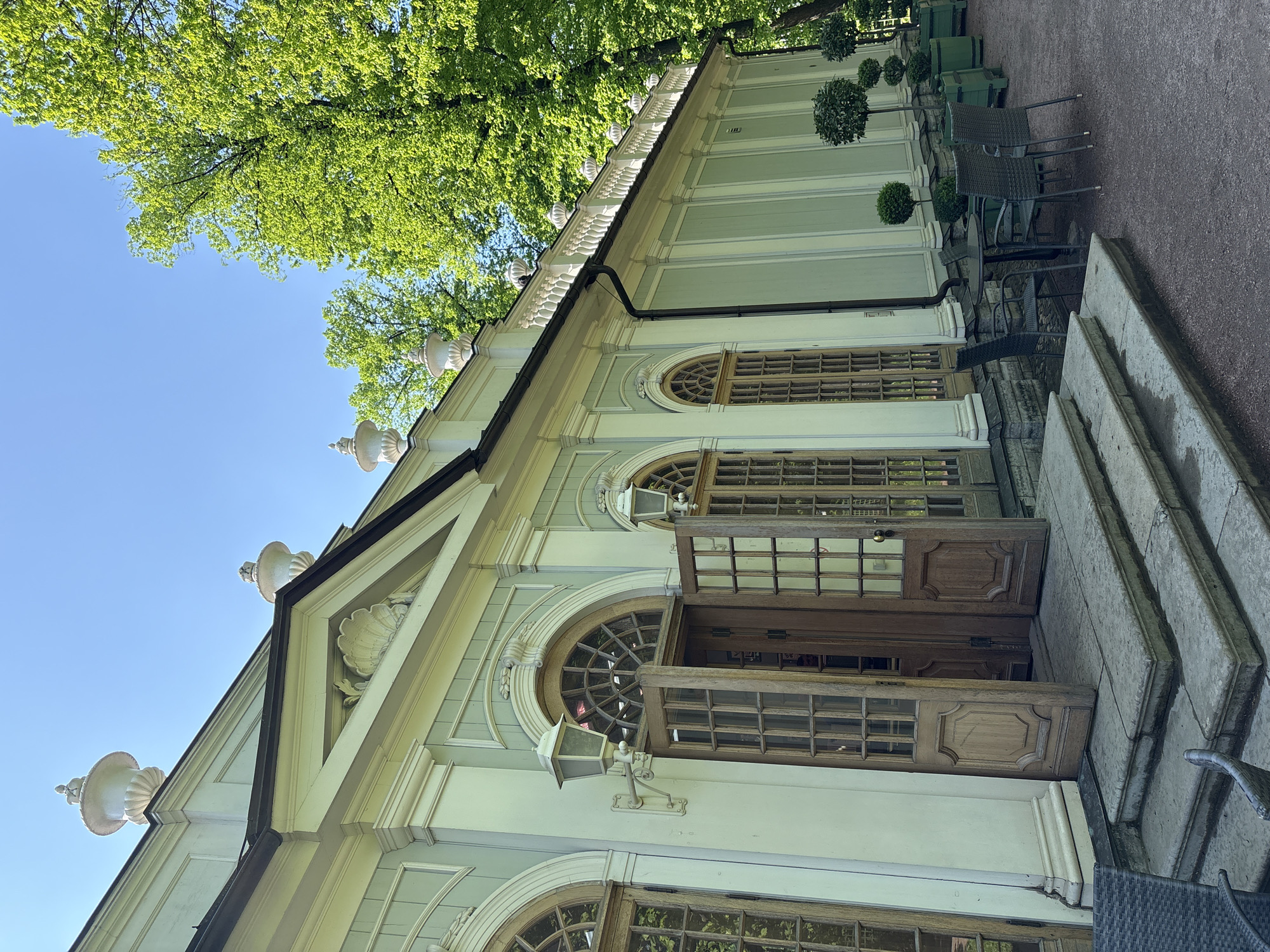 A low-angle shot captures a light green building with neoclassical architectural elements on a sunny day. The structure has a steeply pitched roof adorned with ornamental urns and carved details. Large arched windows, some with open wooden doors, line the building's facade. A set of stone steps leads to the entrance. Outdoor seating with metal chairs and tables is visible to the right of the building. Lush green trees fill the upper part of the frame against a clear blue sky. This is the Cameron Gallery located in the Catherine Park of Tsarskoye Selo, a former imperial residence in the city of Saint Petersburg, Russia.