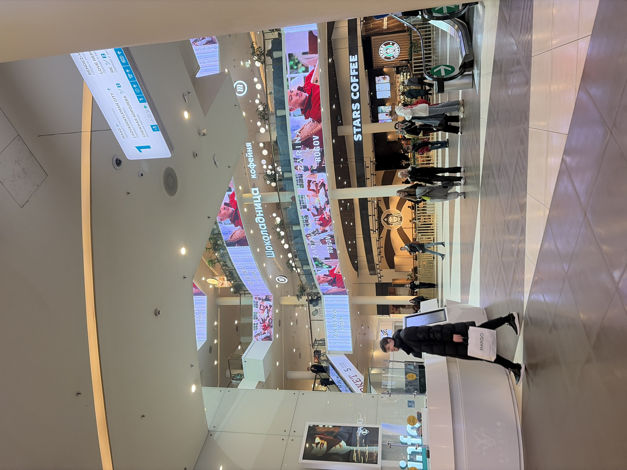 The photograph captures an interior view of a shopping mall, likely in Saint Petersburg, Russia, based on the Cyrillic script and brand names visible. Dominating the upper part of the frame are several large digital display screens that feature advertisements and branding, including the name "ZARINA ROGOV" and "STARS COFFEE" (a replacement for Starbucks in Russia). The screens curve and follow the mall's architectural design.
Below the displays, shoppers are seen walking or standing in line at STARS COFFEE. The architecture of the mall includes vertical support columns and multiple levels. Signage directing customers to different stores, such as LADY&GENTLEMAN CITY, RENDEZ-V, and LOVE REP, is visible on a hanging sign. On the right side of the image, an escalator is seen.
In the foreground, a woman in a black coat is seen standing near a shop named "MANGO" holding a shopping bag from the same brand. The lighting is bright and diffuse, illuminating the scene. The flooring appears to be tiled and a neutral color. The overall impression is of a modern, bustling commercial environment.