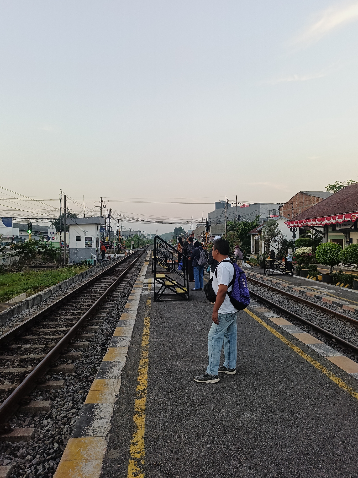 This image depicts a train station platform in Gedangan, Indonesia. In the foreground, a man wearing a white t-shirt, blue jeans, and a purple backpack stands facing away from the camera, looking towards the train tracks. He is positioned on the dark asphalt platform, which is marked with yellow and black safety lines.
Several other people are visible on the platform, some queuing, others walking. They are dressed in casual clothing. Various structures are present, including a small building with a white facade and grey accents, likely a station office or control room, and a longer building with a red and white awning, possibly a waiting area or shop. Train tracks run alongside the platform, with ballast and railway sleepers clearly visible. Overhead power lines and utility poles are present in the background, suggesting an active railway line. Buildings and greenery are visible beyond the tracks and platform. The sky is overcast with a pale, soft light, indicating it may be early morning or late afternoon. The overall scene is a common view of daily life at a local train station.