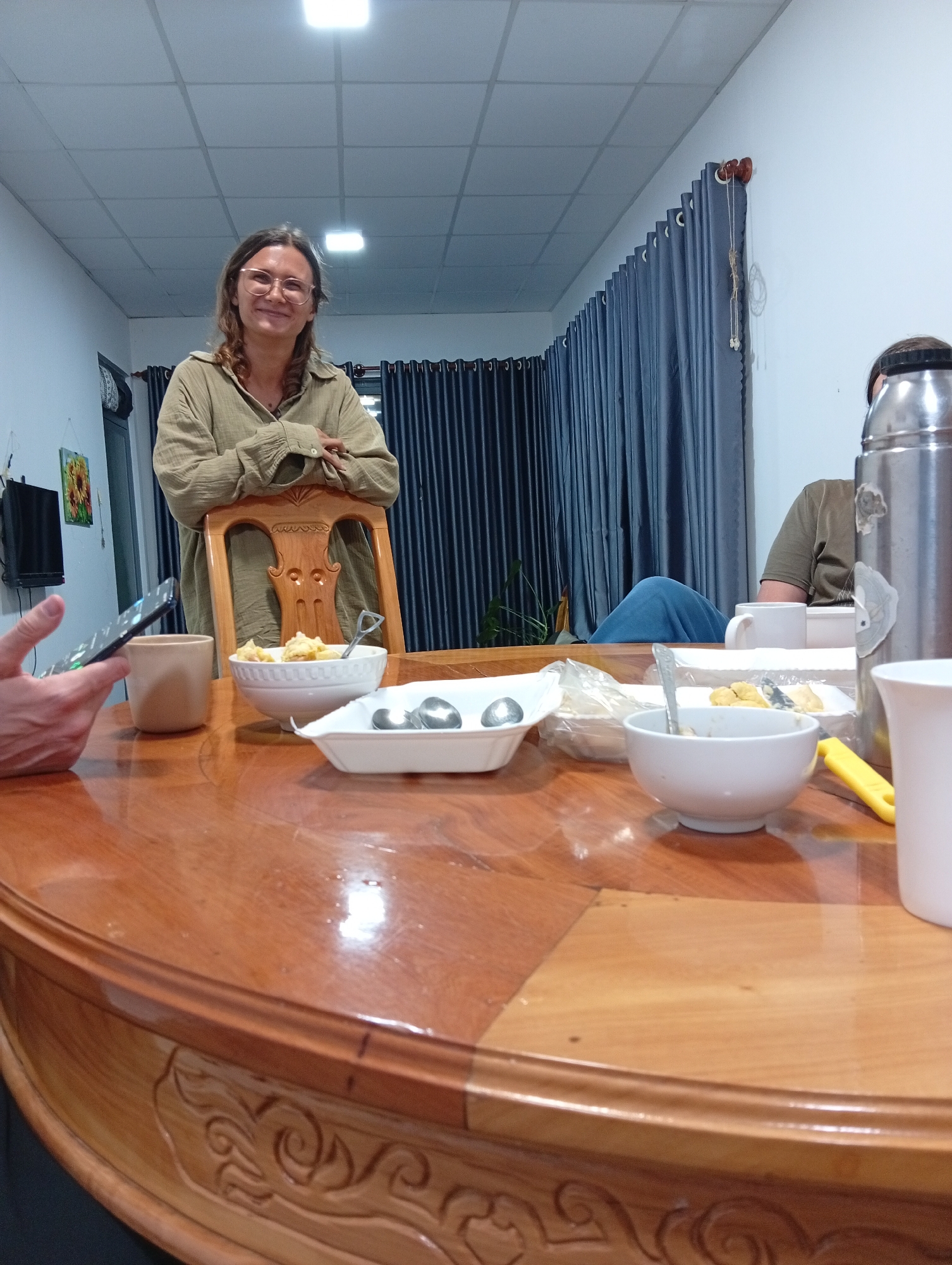 The image captures an indoor dining scene in what appears to be a residence.
People: A woman with glasses leans on the back of a wooden chair. Other individuals are partially visible at the table, including a person holding a smartphone and another wearing a green t-shirt with blue pants.
Subjects/Objects: A large, ornate wooden table dominates the foreground, holding several bowls of food, serving spoons, a coffee mug, a thermos, and a knife. A television and a painting hanging on the wall are in the background.
Environment: The room is brightly lit by ceiling lights. Blue curtains cover a window behind the woman. The location is possibly a home or guesthouse in Da Lat, Vietnam.
Interactions: People are gathered around the table, with the implication of a meal or gathering. Someone is using a smartphone.