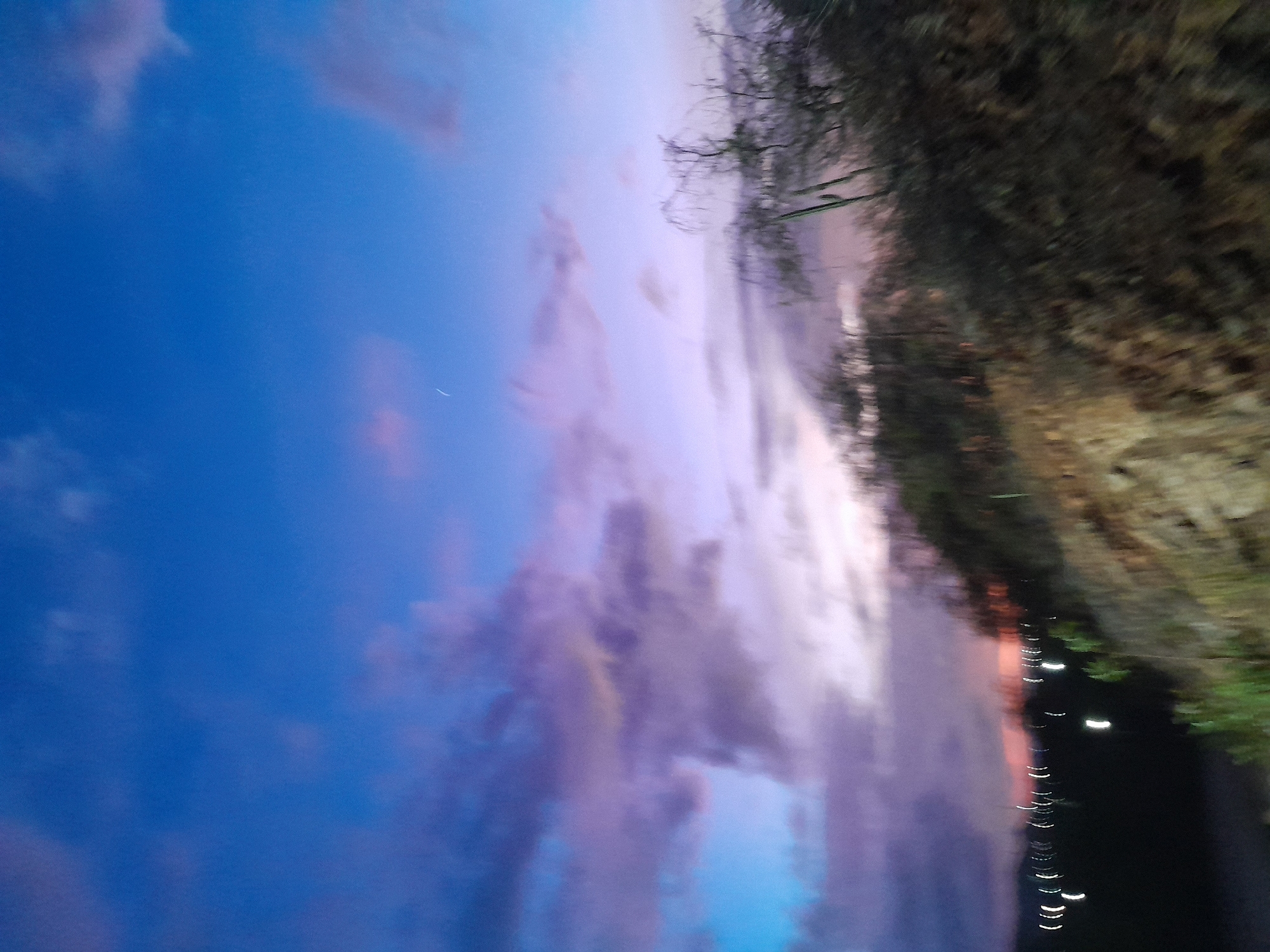 The image captures a blurred twilight scene with a vibrant sky transitioning from deep blue to shades of purple and pink. A few faint clouds are visible, with a thin crescent moon or faint streak of light in the upper portion of the sky. The foreground is dominated by a dark, indistinct landscape of trees and brush, with hints of dry, rocky terrain. In the lower left of the image, a series of blurred white streaks suggest distant lights, possibly from a city or town. The overall impression is one of movement and the fleeting beauty of dusk. The location is identified as Santa Ana, Venezuela, suggesting this scene is from that region.