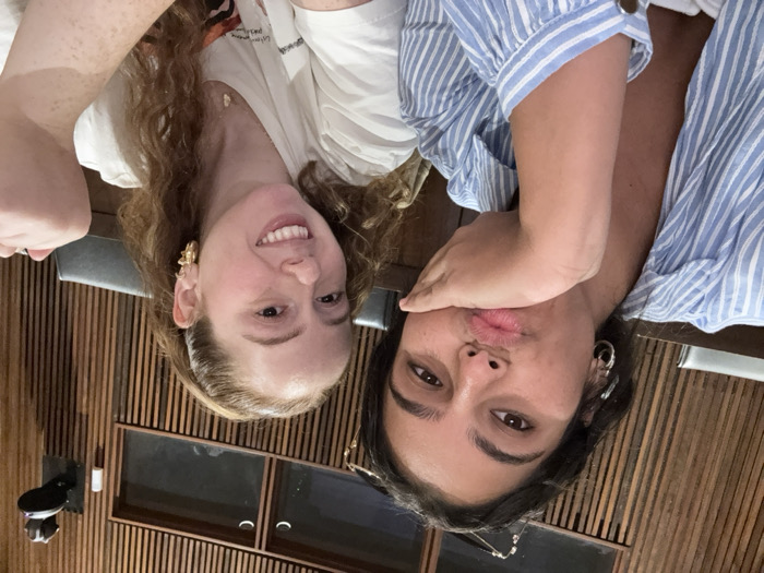A close-up image depicts two young adult females indoors. The woman on the left has dark hair, wears a light blue and white striped button-down shirt with the collar open, and silver hoop earrings. She is making a "duck face" expression with puckered lips and her right hand rests against her cheek. The woman on the right has light brown, curly hair tied back, wears a white short-sleeved t-shirt with a graphic design partially visible, and gold-toned hoop earrings and a delicate necklace. She is smiling broadly. Her left arm is raised with the hand mostly out of frame.
The background features dark wooden walls with vertical slats. In the upper left background, two small, rectangular window-like openings or cutouts are visible. In the upper right, a dark-colored camera or recording device is mounted on the wall. Dark seating is partially visible behind the individuals. The setting appears to be an interior academic space, such as a lecture hall or seminar room, consistent with facilities found within Princeton University in Princeton, New Jersey, United States.