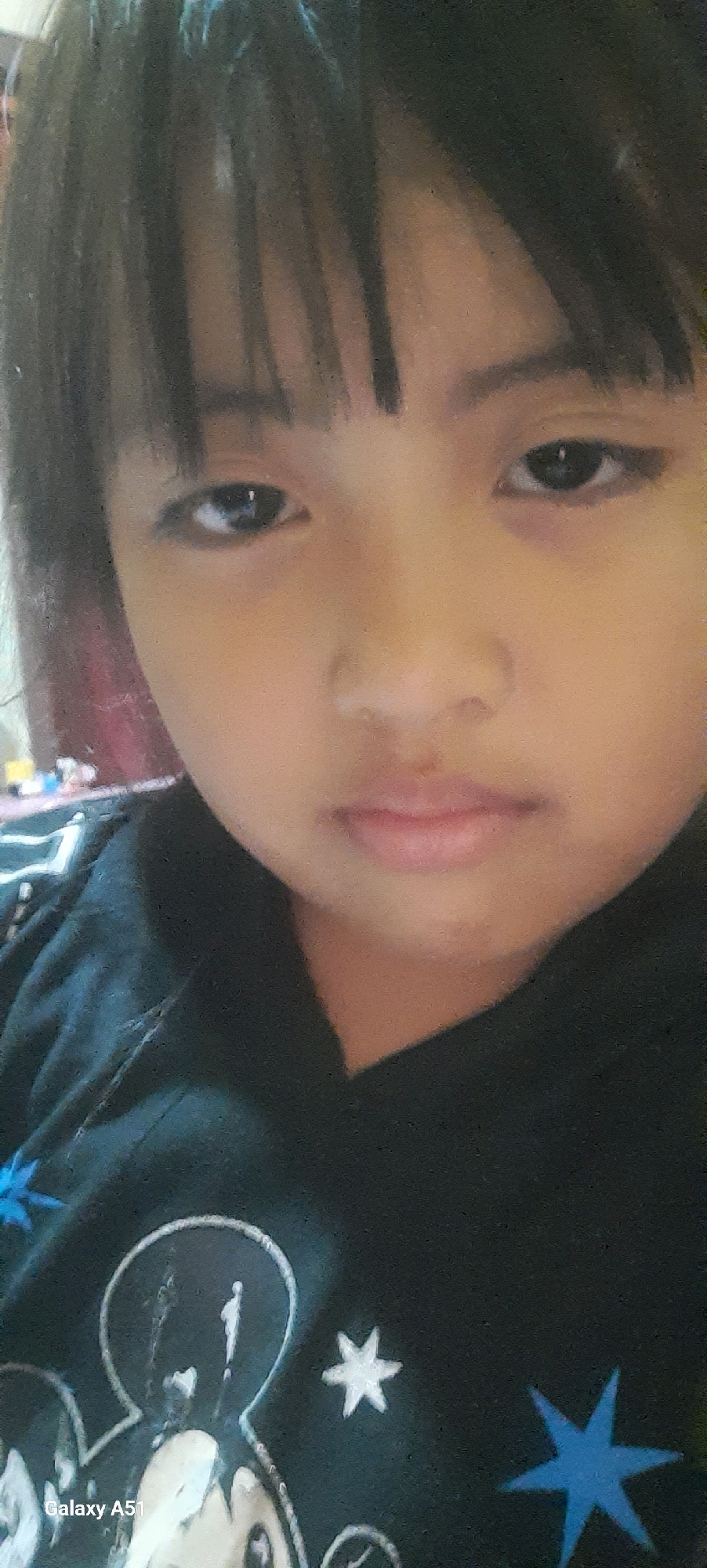 A close-up, low-angle selfie shows a young child with dark hair and bangs, looking directly at the camera with a neutral expression. The child is wearing a black hoodie with a Mickey Mouse graphic and stars. The background is blurred, but appears to be an indoor setting with some colorful objects visible. The bottom left corner of the image shows the text "Galaxy A51". The child's face is the primary focus of the image, with natural lighting illuminating their features.