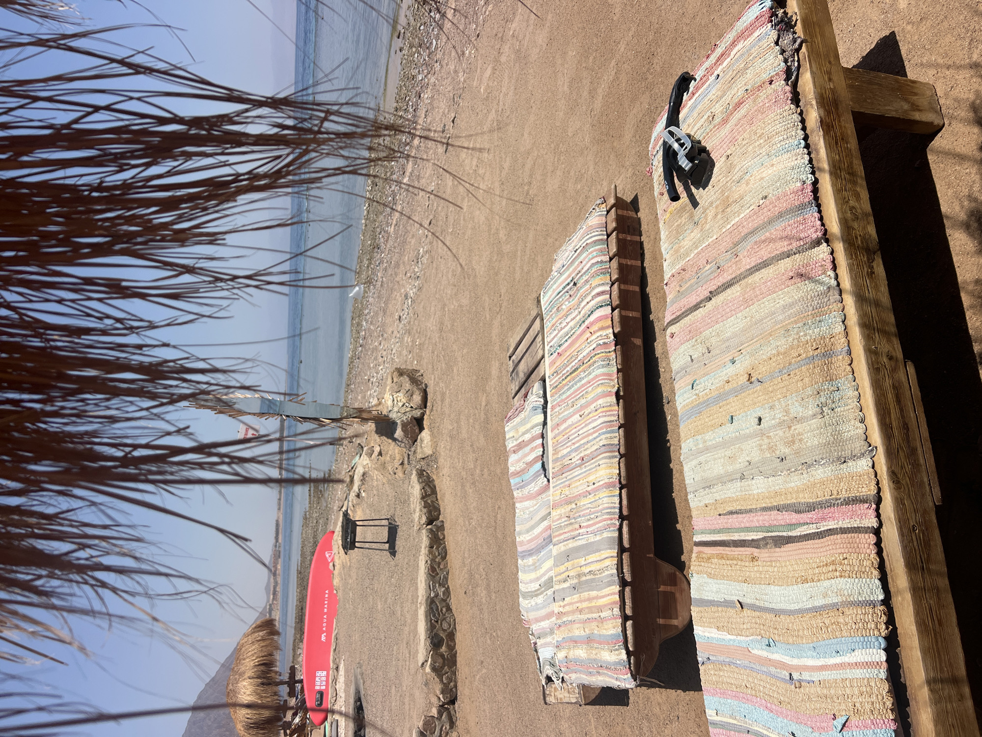 Two wooden sun loungers with colorful striped mats are positioned on a sandy beach in Nuweiba, Egypt. The sun loungers are oriented towards the calm, blue sea, which is visible in the background. Behind the loungers, there is a beach umbrella and a red surfboard leaning against it. Further back, the shoreline is lined with small stones and pebbles, and the rugged mountains of the Sinai Peninsula are visible in the distance under a clear, bright sky. The scene is framed by the dangling fronds of a palm tree in the foreground, casting dappled shadows on the sand.