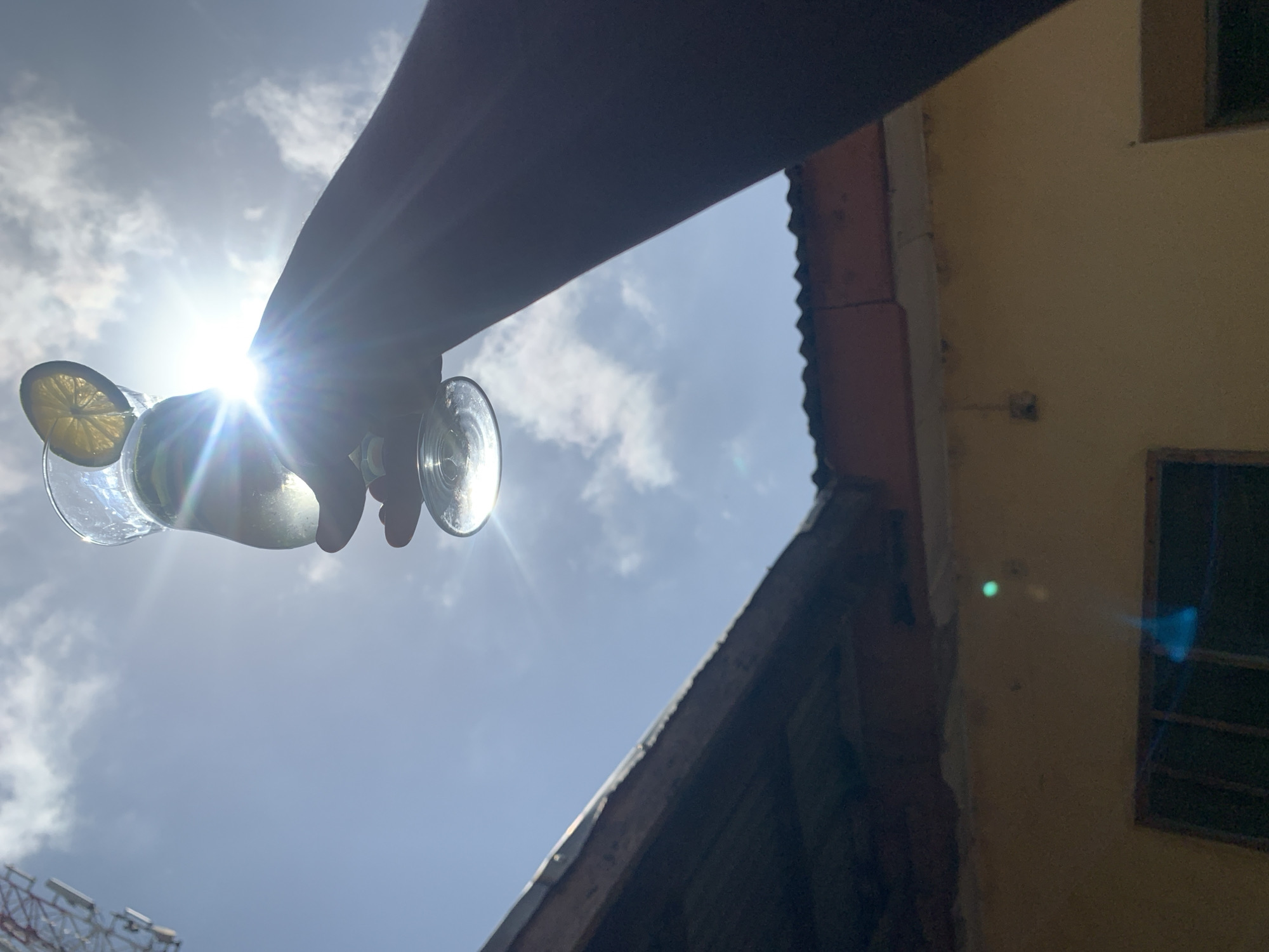 A hand holding a glass with a lemon slice against a bright sky with scattered clouds, partially obscuring the sun. A portion of a building is visible on the right, with windows and a slightly dilapidated exterior. A structure, possibly a tower, is faintly visible in the lower left corner. The city of Rafin Par, Nigeria, cannot be definitively determined from the image.