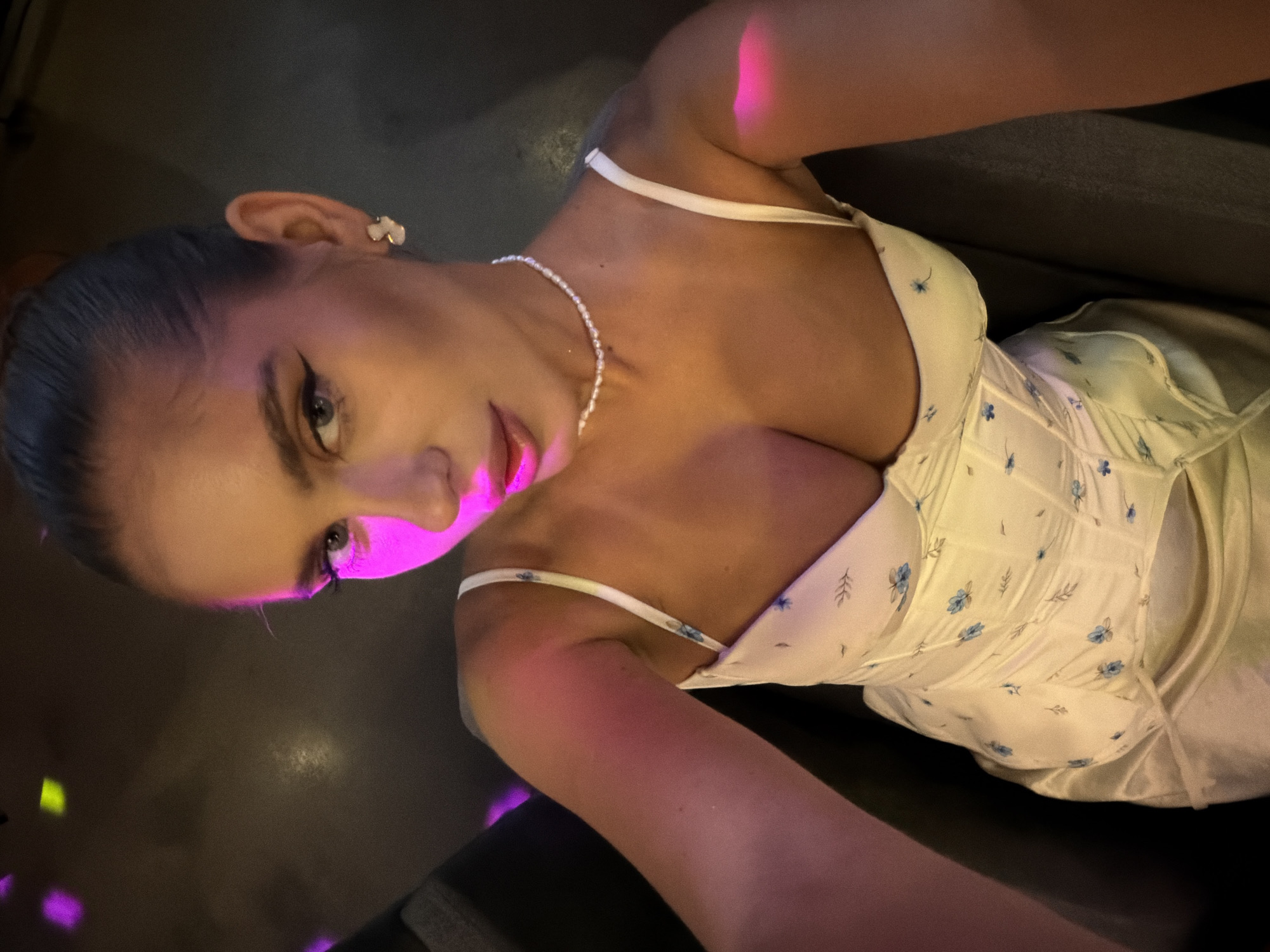 A high-angle, close-up selfie taken from the chest up shows a young woman with dark hair pulled back. She is wearing a white slip dress with a floral pattern and a pearl necklace. The lighting is dim and illuminated by pink and purple lights, creating a moody atmosphere. The background is dark and out of focus, suggesting an indoor setting, possibly a club or bar in Krasnoyarsk, Russia.