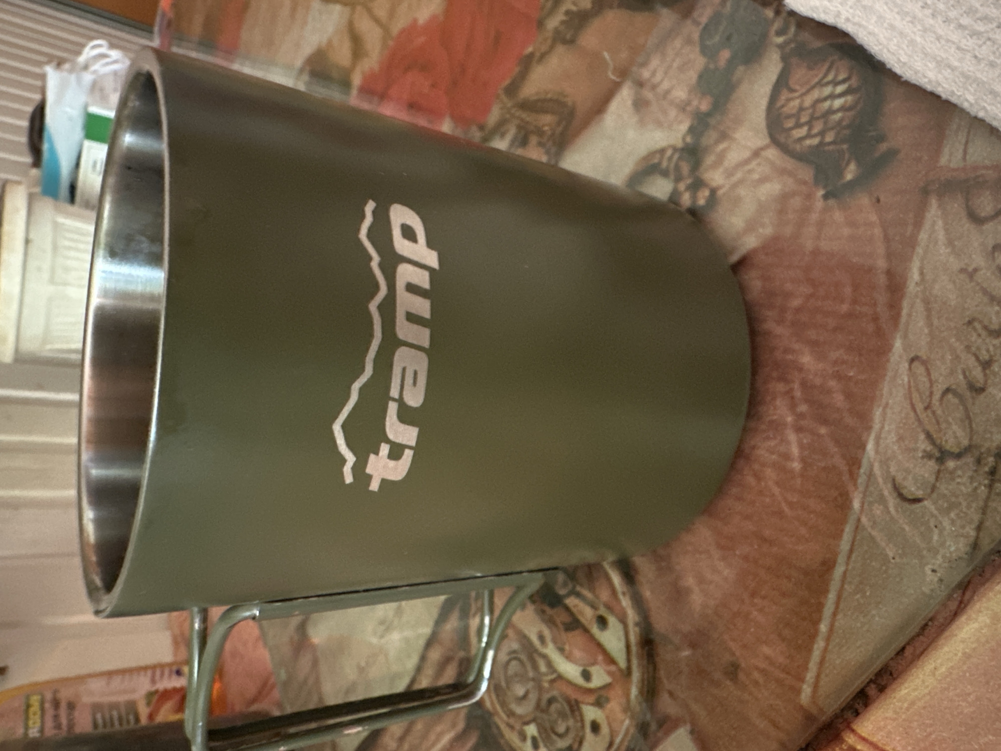 A green metal mug with a silver handle and rim sits on a table covered with a patterned tablecloth. The mug has a white logo on it that appears to be mountains with the word "tramp" underneath. The background is blurred, showing a wall and some shelves with items on them. The overall scene is an indoor setting, likely a home or restaurant. The image does not provide any information or context about the city of Podilsk, Ukraine.