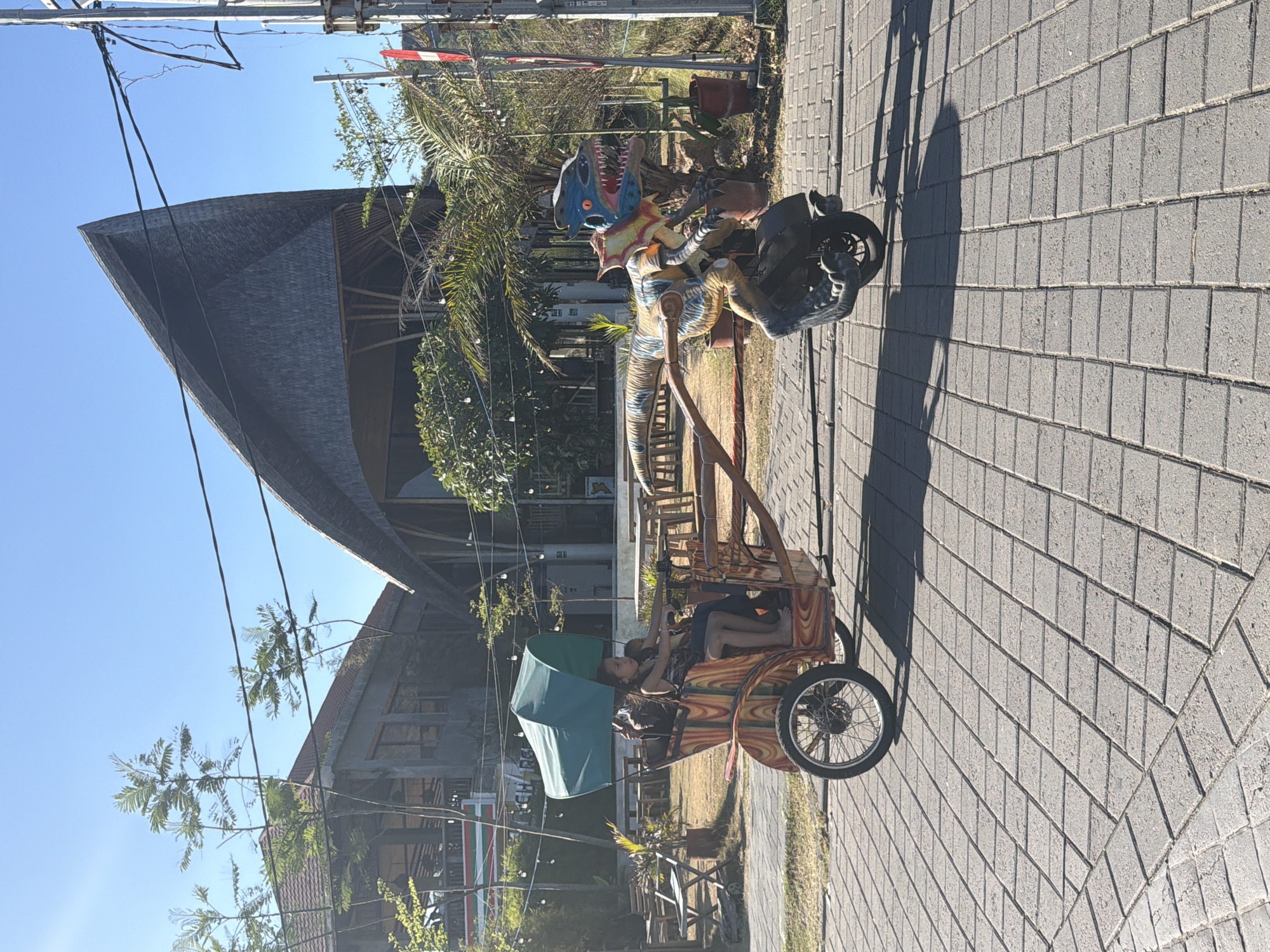 The image captures a sunny day scene in Kuta, Indonesia. The central focus is a **tricycle carriage** with wooden construction, adorned with colorful patterns. Two **people**, likely children or young teenagers, are seated inside the carriage, one holding onto the frame. A green **umbrella** provides shade over them. The carriage is parked on a **paved walkway** made of gray interlocking bricks.
In the background, a building with a **thatched roof** and a distinct triangular shape is visible, suggesting a resort or restaurant setting. Another building with a reddish-brown roof is also present. A **dinosaur statue** mounted on what appears to be a motorcycle is positioned to the left of the walkway, adding a playful element to the scene. Palm trees and other lush **vegetation** are scattered around, characteristic of a tropical environment. Power lines are strung across the top of the frame. The lighting indicates it is daytime, with clear blue skies suggesting fair weather. The overall mood appears relaxed and pleasant. No visible text is discernible in the image.