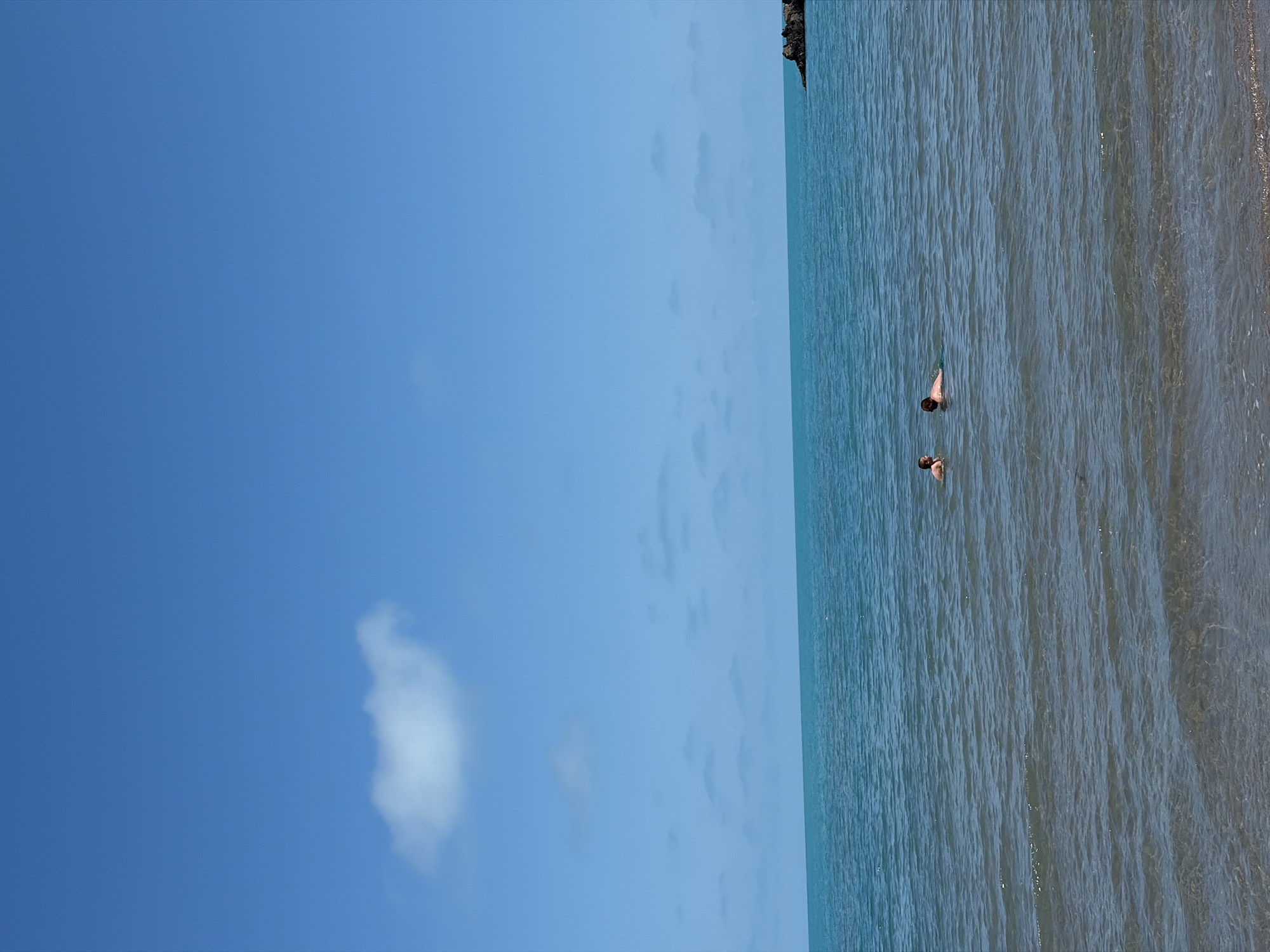 Two individuals are partially submerged in the clear, turquoise waters of the Black Sea at Rezovo, Bulgaria. The person on the left, appearing smaller, is facing towards the right, with their head and shoulders visible above the water. To their right, the second individual is also facing right, their head and upper torso visible, and they are in motion, possibly swimming or wading. The water is calm with gentle ripples, and the seabed is visible beneath the surface in shallower areas. The horizon is clearly defined, separating the sea from a bright blue sky dotted with scattered, wispy white clouds. In the far distance to the right, a dark, rocky outcrop juts out of the water. The overall scene conveys a serene and warm day at the beach.