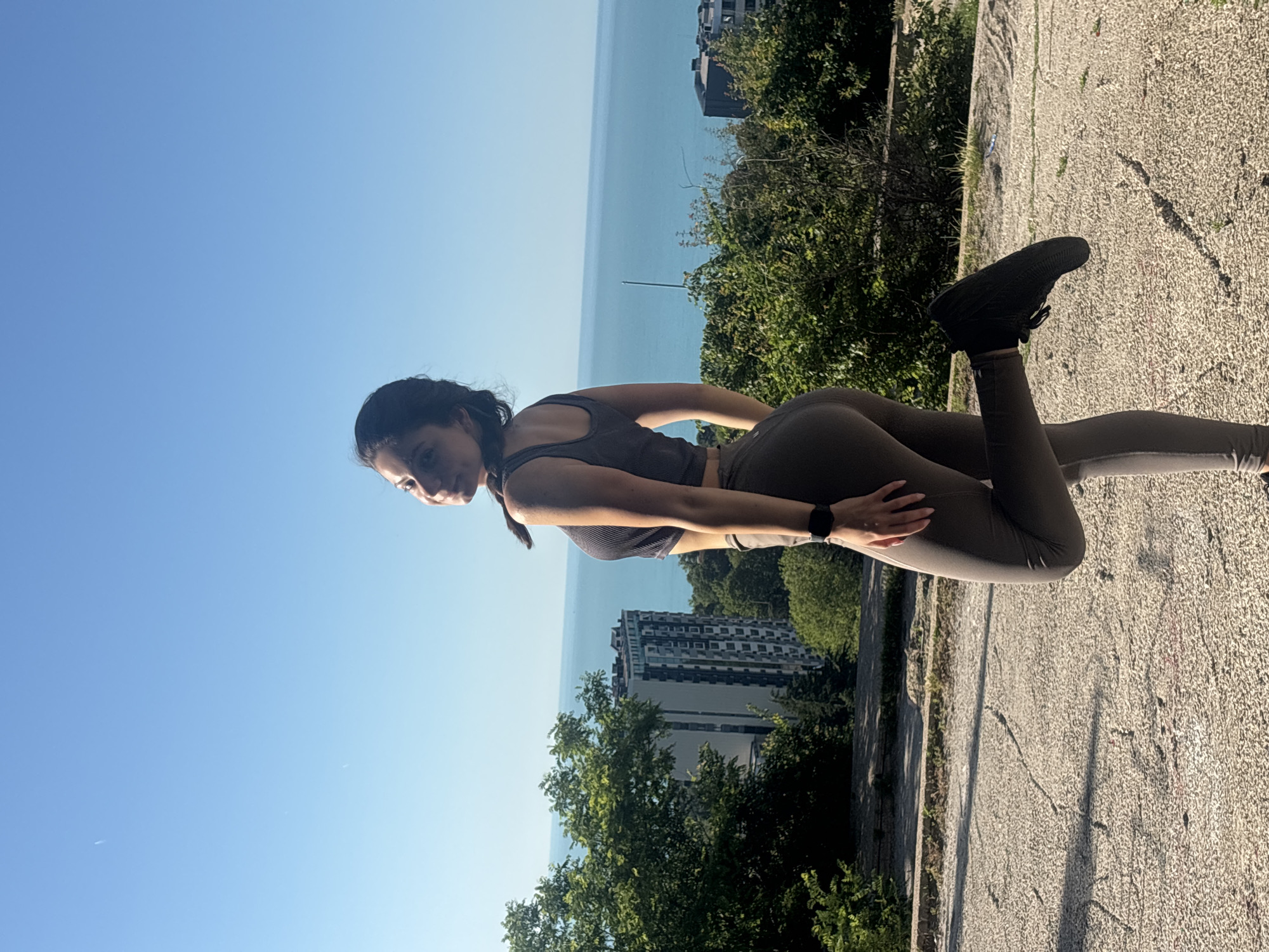 In the city of Varna, Bulgaria, a young woman is photographed outdoors. She is standing on a cracked concrete pathway with lush green trees and buildings in the background, with a view of the Black Sea. The woman is wearing a dark gray sleeveless top and matching leggings, and black athletic shoes. Her hair is dark and styled in a braid. She is posing with one leg bent at the knee and her arm resting on her thigh. The sky is a clear, bright blue. The sun appears to be shining, casting shadows on the ground.