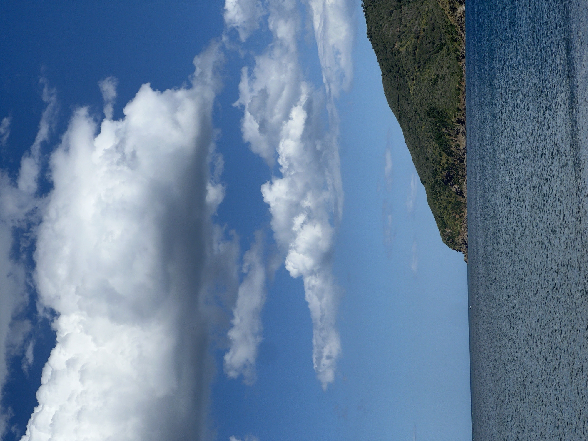 The image shows a view of M'Diq ⵎⴹⵢⴰⵇ المضيق, Morocco. The right portion of the image is dominated by the sea. Adjacent to the sea is a steep, green, mountainous coastline. The left portion of the image contains a blue sky and fluffy, white clouds.