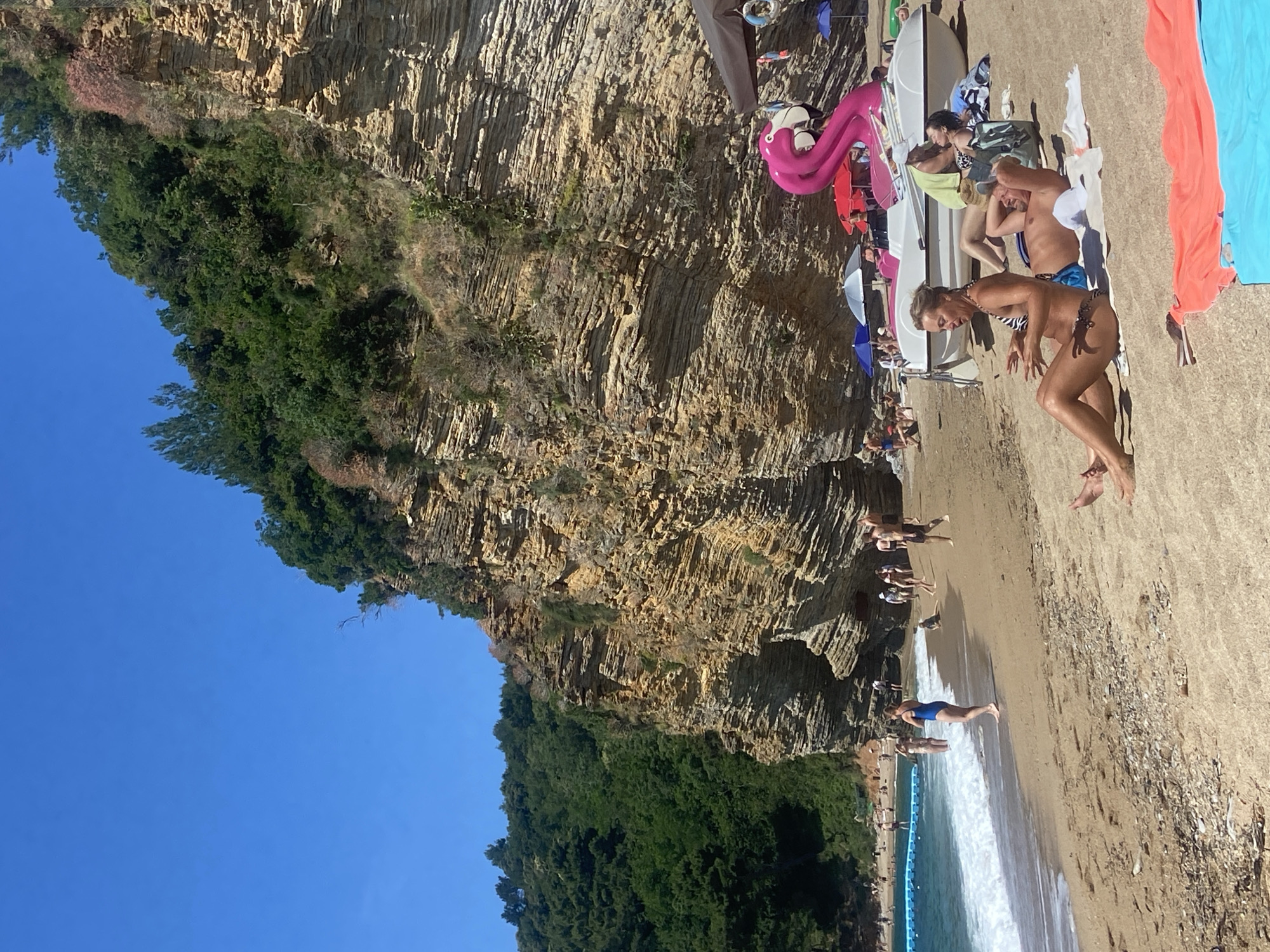 The image depicts a lively beach scene in Sveti Stefan, Montenegro, set against a stunning natural backdrop under a bright blue sky. A towering cliff face, characterized by distinct horizontal layers of rock, rises dramatically on the left side of the frame, partially covered by lush green vegetation.
On the sandy-pebbly beach, numerous people are enjoying a sunny day. In the foreground, a woman with light hair, wearing a black and white striped bikini, is captured in a dynamic, leaning pose near the water's edge, seemingly engaged in an activity or about to enter the sea. Beside her, a man in blue swimming trunks stands facing the water. Further back, more individuals are relaxing on towels, with some gathered around white boats or large paddleboards. A prominent pink inflatable flamingo, colorful towels (orange and blue), and several beach umbrellas add vibrant touches to the scene.
The calm sea, visible on the right, features gentle waves breaking near the shore, with a blue floating barrier in the distance marking a designated swimming area. The bright sunshine and clear skies suggest it is likely mid-day, creating clear shadows and a pleasant atmosphere for beachgoers. There is no visible text in the image.