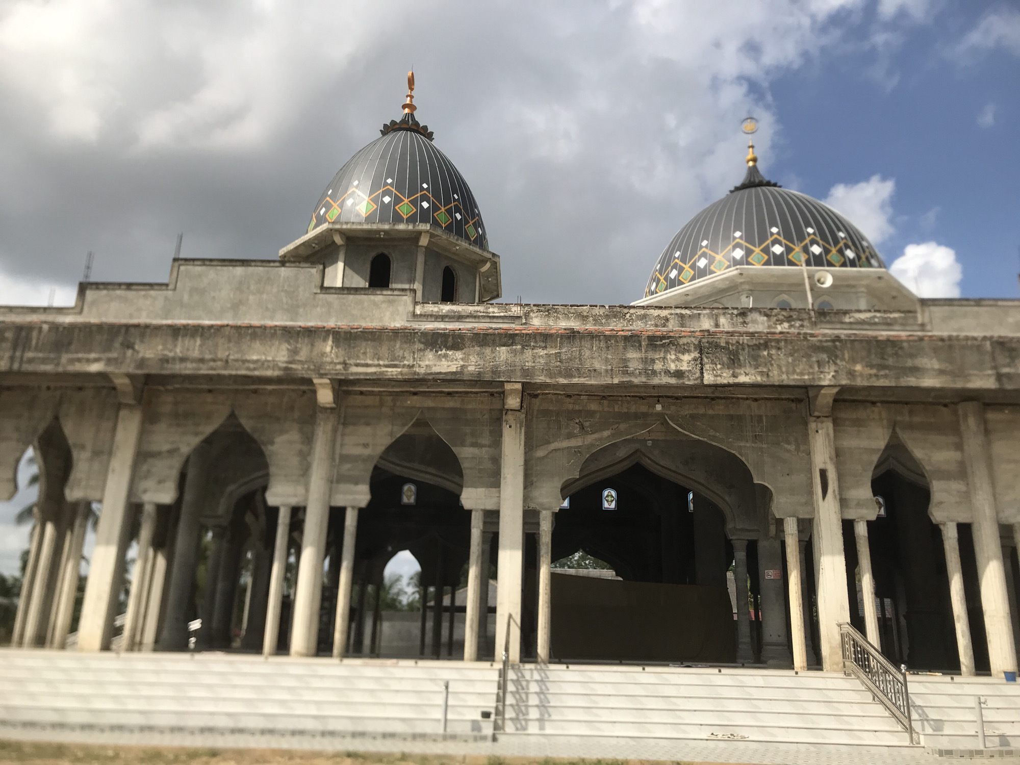 The image shows the front facade of a mosque in Nicahawe, Indonesia. The building is constructed of light-colored concrete and features two large, dark gray domes decorated with geometric patterns in white, green, and yellow. Each dome is topped with a spire and a crescent moon symbol.
The mosque has a series of arches supported by columns, creating a covered walkway. Stairs lead up to the main entrance. The building appears unfinished, with exposed concrete and visible construction elements.
The sky behind the building is partly cloudy, with patches of blue visible. The foreground is a grassy area. The lighting is bright, suggesting a daytime scene. There are no people visible in the image.