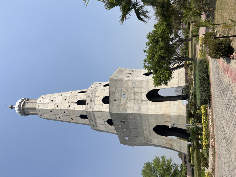 The image shows a tall, concrete tower with a domed roof. The tower is located in a park, with green lawns and trees surrounding it. The tower has a series of arches and windows, and the concrete is a light gray color. The tower is a historical monument called the "Shaheed Smarak" located in Sahibzada Ajit Singh Nagar, Punjab, India. The tower is dedicated to the memory of those who died fighting for freedom. The tower stands tall against a clear blue sky, and there are no people present in the scene. The ground is paved with a gray brick pattern and a small red brick pattern. The image is taken from a low angle, looking up at the tower. The overall scene is peaceful and serene.