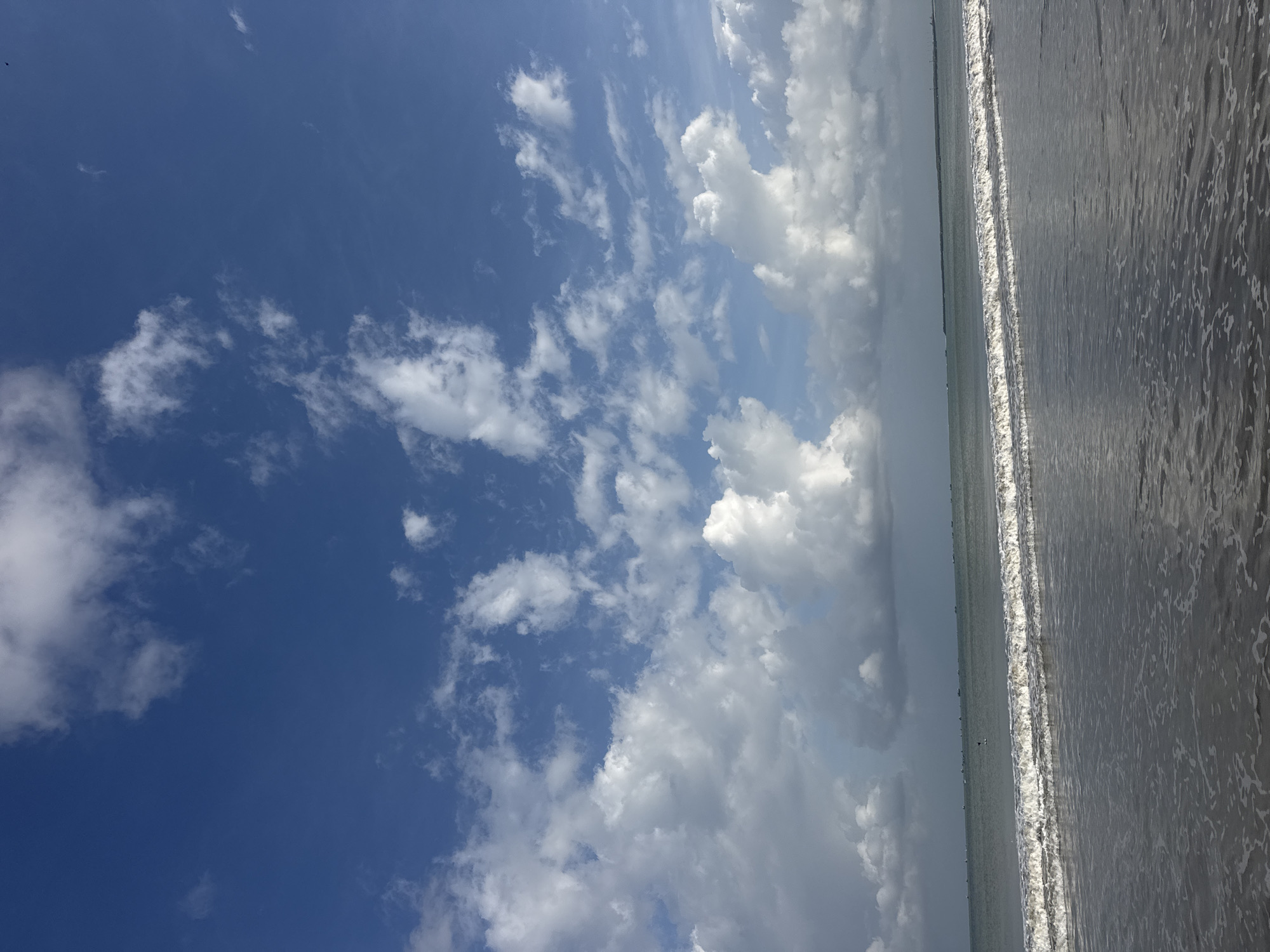 The image depicts a serene beach scene on a partly cloudy day, likely in Kuta Selatan, Indonesia. The foreground is dominated by the ocean's edge, with gentle waves lapping at the sandy shore. The water is a mix of murky grey and foamy white where the waves break. In the mid-ground, a line of calm sea stretches towards the horizon, where a faint, dark line suggests a distant landmass or breakwater. Along this line, several small, dark shapes, possibly boats, are visible. The sky above is a vibrant blue, dotted with scattered white cumulus clouds, some of which appear fluffy and well-defined, while others are more wispy and diffused. The overall atmosphere is one of tranquility and natural beauty.