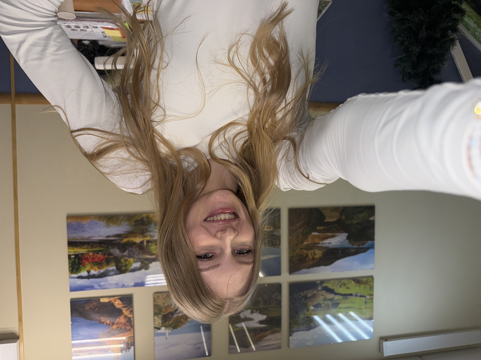 A blonde woman in a white long-sleeved shirt is smiling into the camera, which is being held upside down. Behind her, there are six landscape paintings hung on a beige wall. The paintings depict bodies of water, mountains, and green fields. A white rectangular object, potentially a light fixture, is situated on the bottom right. The ceiling is a dark blue-grey, and there is a leafy green plant in the top right corner. There is no visible information to determine the location as Perm, Russia.