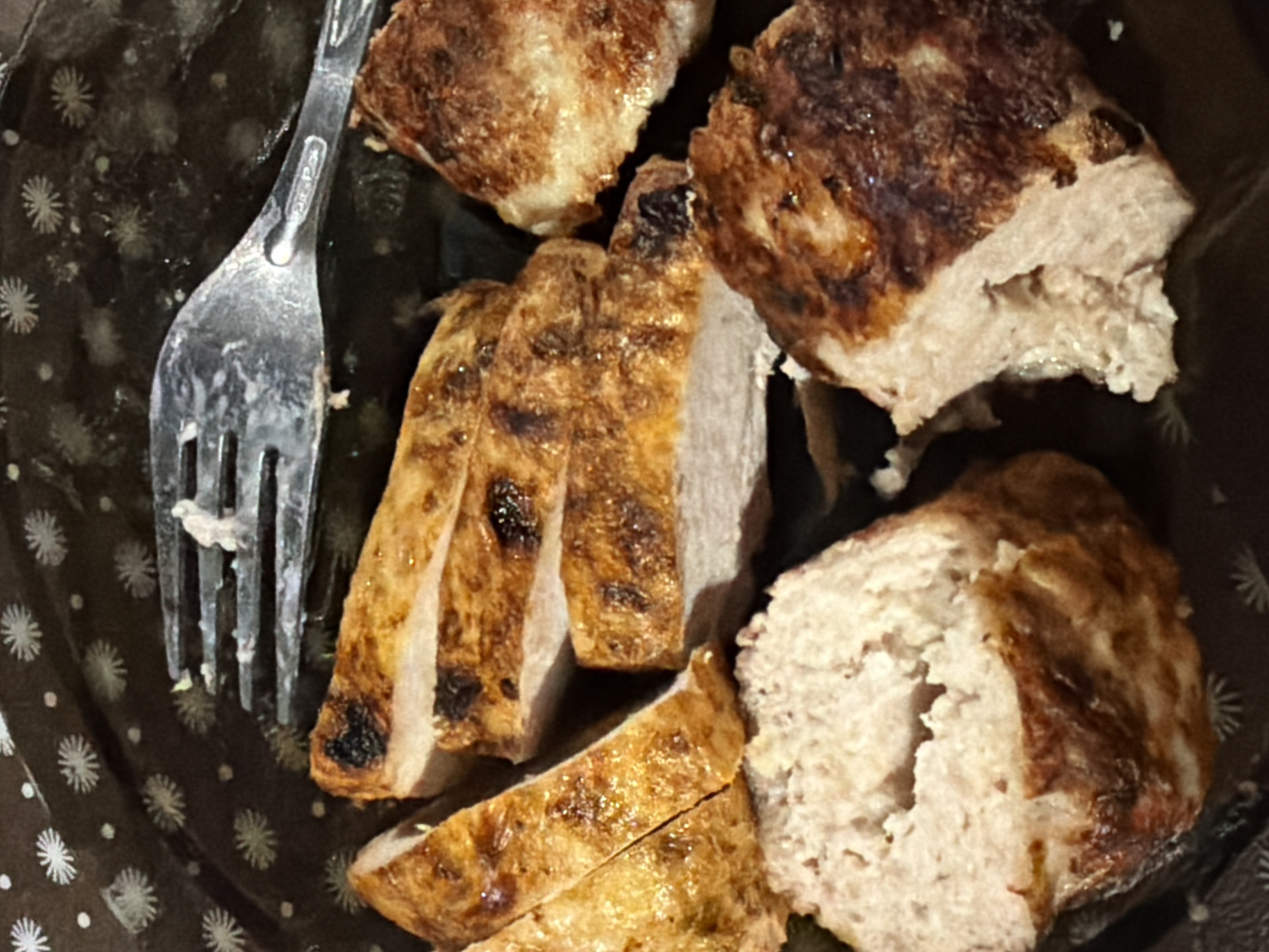 The image shows a close-up of several pieces of cooked meat, likely chicken or pork, on a dark, patterned plate. The meat appears to be sliced and has grill marks. A fork is visible in the upper right quadrant of the image, with a small piece of meat on it. The background of the plate has a repeating pattern of white starbursts or flowers. The lighting suggests an indoor setting, possibly a restaurant or home. There is no visible indication of the city of Sochi, Russia, in the image.