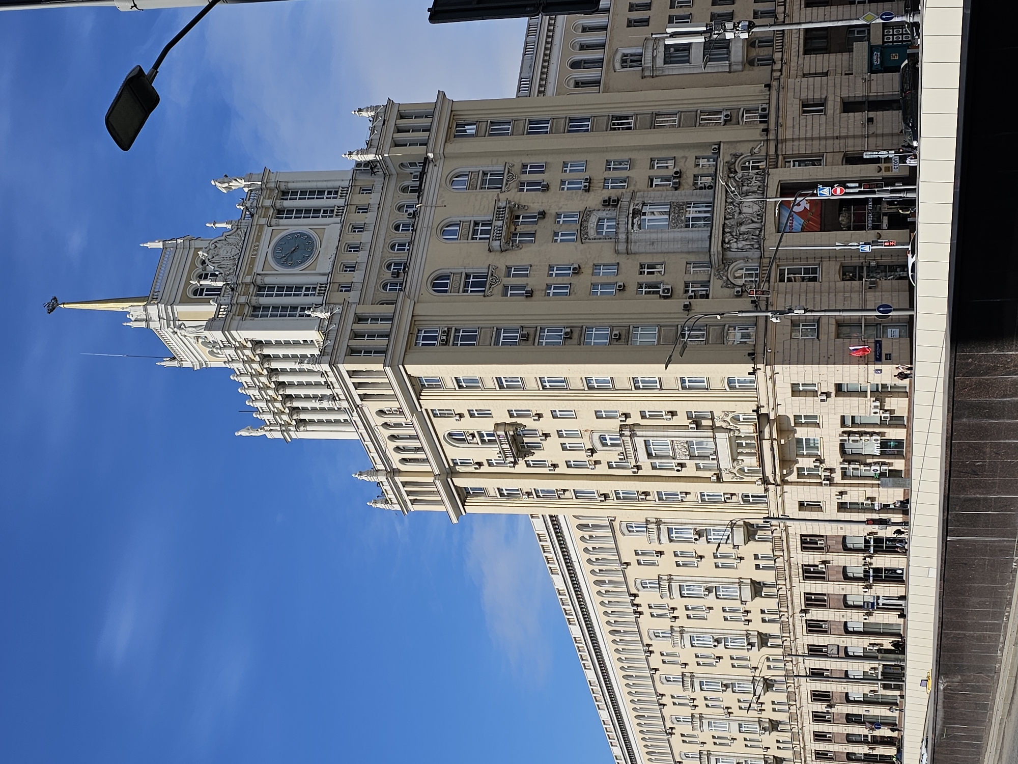 Here is a description of the image:
The image depicts the Kotelnicheskaya Embankment Building, one of the Seven Sisters skyscrapers in Moscow, Russia. The building is prominently featured against a bright blue sky with a few wispy clouds. Its architectural features, including its clock tower, spire, ornate facade, and multiple windows, are clearly visible. Street lighting fixtures are partially visible in the foreground. The building is shown from a low-angle perspective, emphasizing its height. There is a street with a sidewalk on the right side of the building and traffic lights on the street.