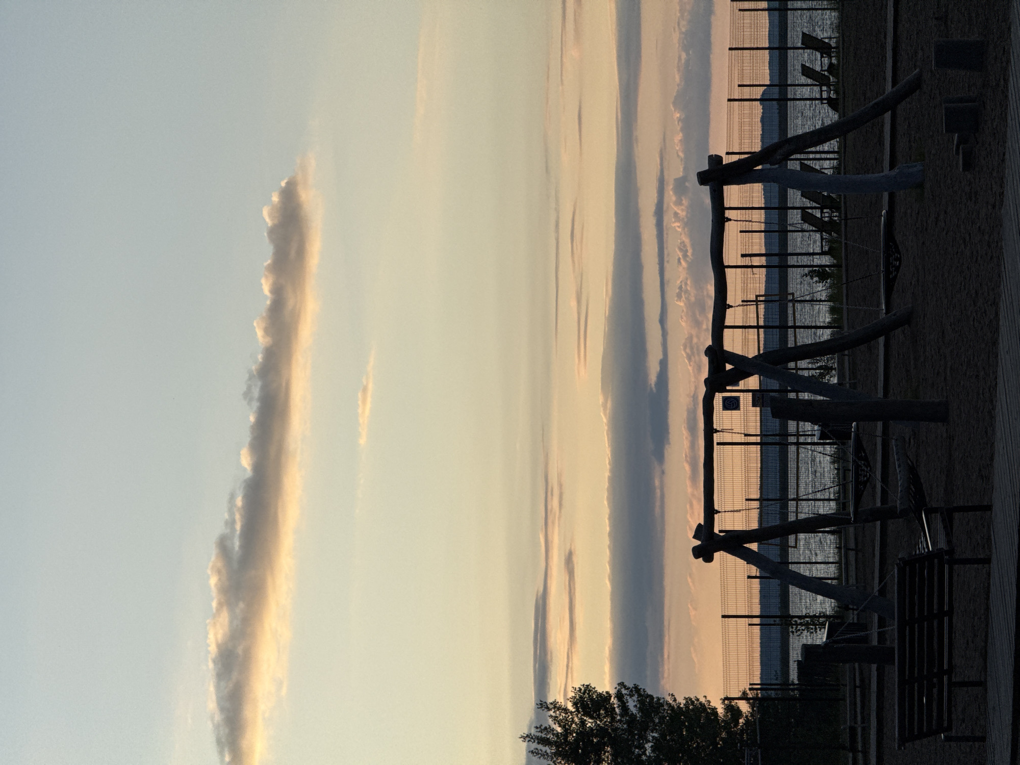 This media file presents a serene outdoor scene captured during sunset or the golden hour, looking out over a body of water from a recreational area in Chaikovsky, Russia.
In the foreground, silhouetted against the vibrant sky, stands a playground structure constructed from dark wooden logs, with a swing clearly visible. Dark benches and a ground covering of sand or dirt are also present. A tall chain-link fence runs across the midground, serving as a distinct barrier between the play area and the water. A small sign on the fence displays a white symbol resembling a volleyball.
Beyond the fence, a calm body of water, likely a lake or large river, stretches into the distance, reflecting the warm hues of the sky. A faint, dark outline of distant land or hills marks the horizon. The sky is a striking feature, transitioning from a light, pale blue at the top to warm yellows, oranges, and soft pinks closer to the horizon. Long, wispy clouds are scattered across the sky, catching the golden light and adding textural interest. A dark silhouette of a tree or bushes occupies the lower right portion of the image.
No people are visible, contributing to the tranquil and peaceful atmosphere of the scene. The image evokes a sense of quiet contemplation as day transitions to evening in a scenic waterfront park.