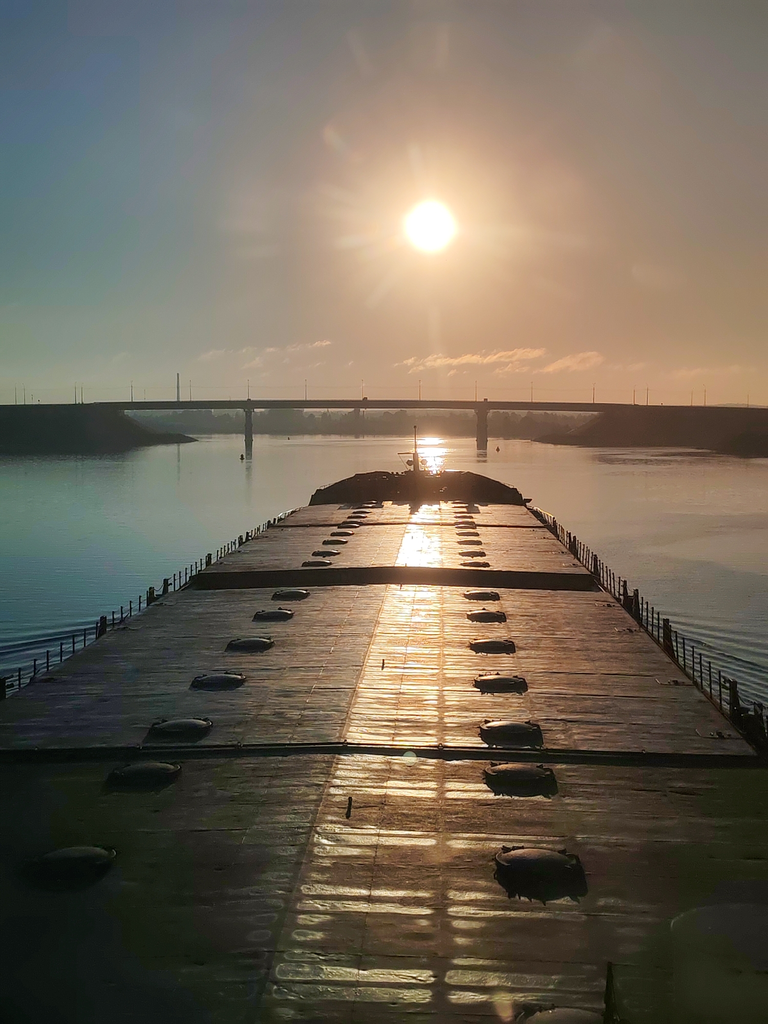 A view from the deck of a barge or ship, looking forward toward a bridge over water. The sun is low on the horizon, creating a strong glare on the water and deck. The deck is flat and appears to be made of planks, with a row of dark, oval-shaped objects running down the center. A metal railing runs along the sides of the deck. The bridge in the distance is a simple, low-slung structure with concrete supports. There are no people visible in the image. The location is unknown.