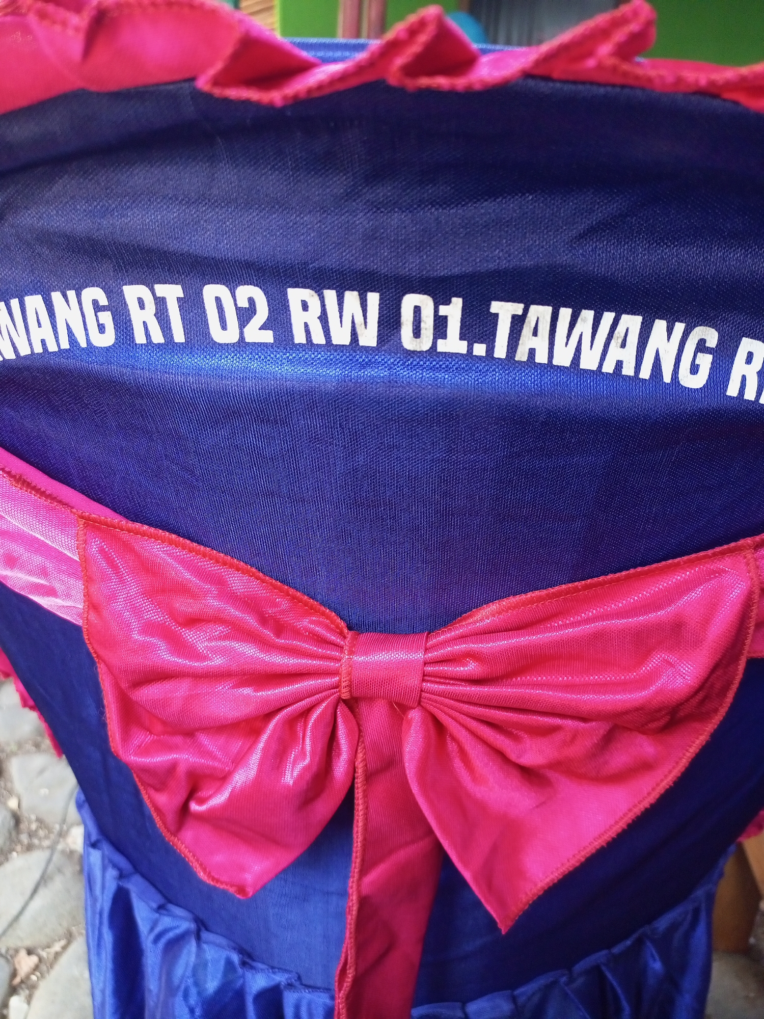 The image shows a close-up of a dark blue fabric, possibly a chair cover or banner, with white text printed on it. The text reads "WANG RT 02 RW 01.TAWANG R". A bright pink ribbon bow is attached to the fabric, positioned below the text. The fabric appears to have a slight sheen and texture. The background is blurred and shows a rough, greyish surface, possibly pavement or cobblestones, and some greenery. The overall impression is of an outdoor setting, though no specific city landmarks are visible, and the location in Jatisrono, Indonesia, is not discernible from the visual information.