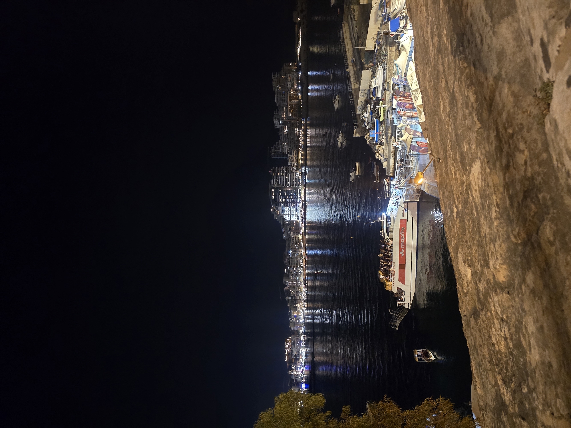 The image captures a night scene in Valletta, Malta, overlooking a harbor. A large white ferry, labeled "mapfire," is docked, carrying numerous passengers on its deck. A smaller, traditional Maltese boat is also visible in the water nearby. The harbor is filled with various other boats, some docked and others moored. In the background, the city skyline is illuminated by numerous lights, with buildings reflecting on the dark water. The foreground features a textured, stone wall, suggesting a viewpoint from a higher elevation, possibly a fortification or terrace. The overall atmosphere is that of a lively evening by the water in a European capital.