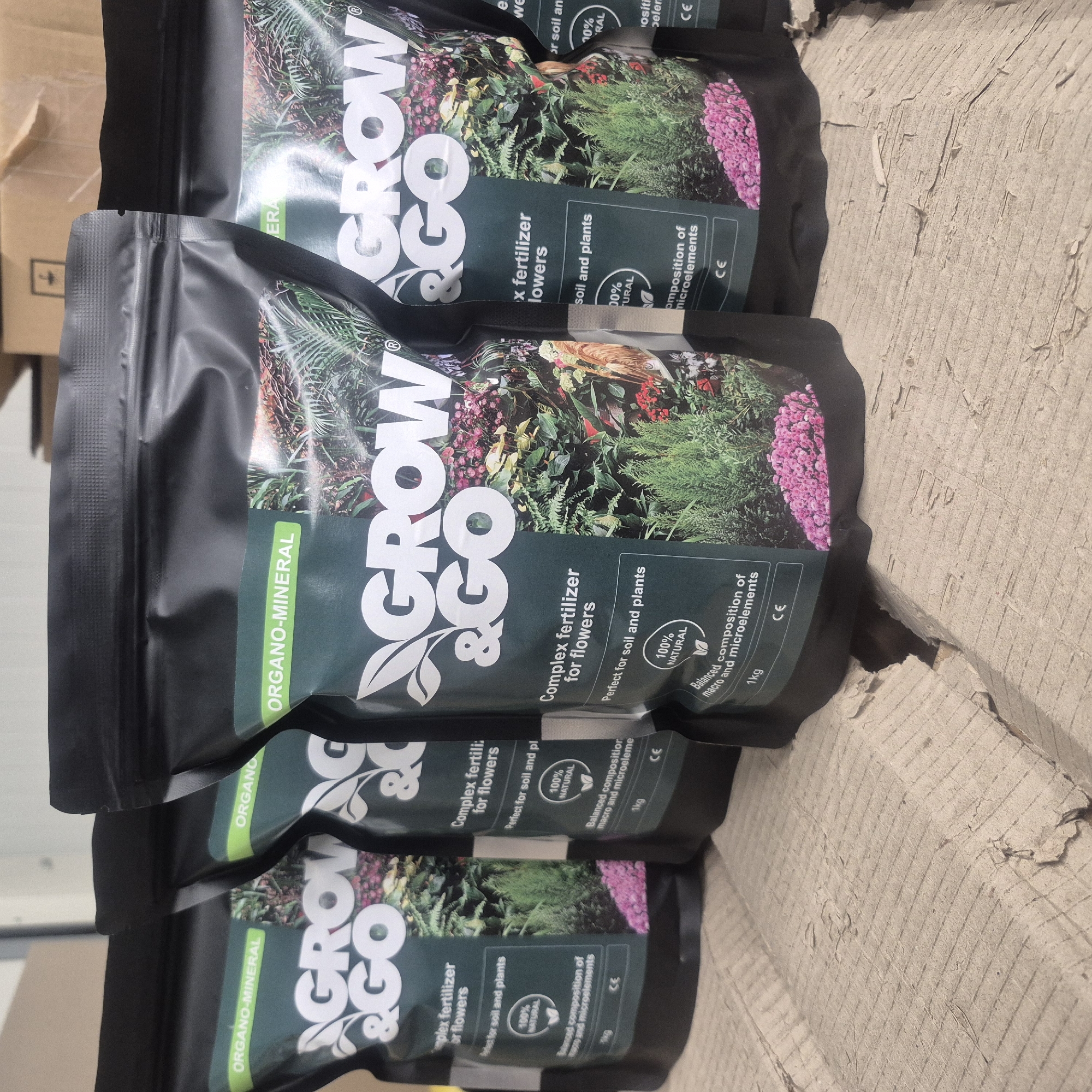This media file shows a stack of at least three bags of "GROW & GO" plant fertilizer. The bags are dark gray with a green label that features images of various plants, including flowers and ferns. The text on the bags indicates they contain "ORGANO-MINERAL Complex fertilizer for flowers," are "Perfect for soil and plants," and have a "100% NATURAL" composition of "macro and microelements." Each bag is labeled as "1kg" and features a "CE" mark. The bags are stacked on a light brown, textured surface that appears to be cardboard or a similar material. In the background, a portion of a cardboard box with a yellow caution symbol can be seen. The lighting suggests an indoor setting, possibly a warehouse or storage area, with no specific time of day or weather conditions discernible. The image is focused on the product packaging, with the bags arranged in a slightly offset, diagonal manner. There are no people or animals visible in the image. The overall impression is of a commercial product display or inventory.