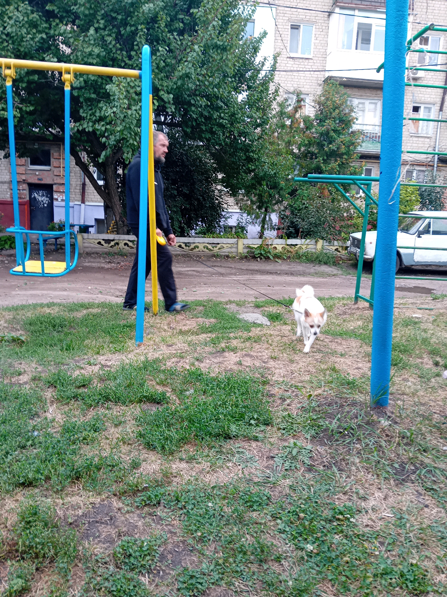 A man is walking a small dog on a leash in a yard with a playground. The man, wearing a black hoodie and dark pants, is on the left side of the frame, looking towards the right. The dog, predominantly white with tan markings, is in the lower right quadrant of the image, walking towards the viewer. A swing set with blue and yellow supports is to the left of the man. In the background, a multi-story brick building is visible, along with trees and a white car. The ground is covered in patches of grass and dirt. The scene appears to be in a residential area of Chuhuiv, Ukraine.