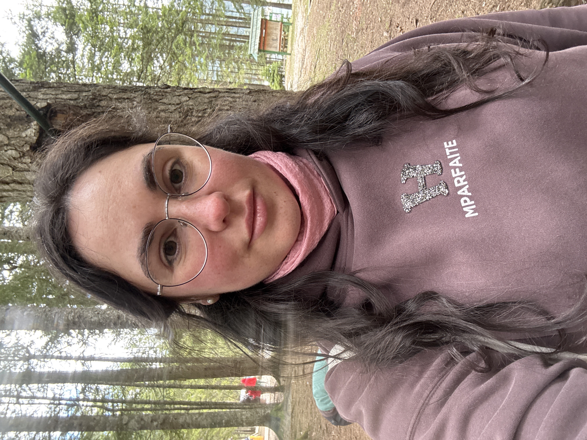 A woman with fair skin, wearing wire-rimmed glasses, a pink neck gaiter, and a mauve hoodie with a logo that reads "HMPARFAITE," is the central subject of this portrait. The woman's long, dark hair has a wavy texture. She appears to be standing outdoors, in a forest setting with tall, slender trees with light-colored trunks. A small, green building with a wooden frame and a sign can be seen in the background, suggesting a park or recreational area. The ground is covered with dirt and leaves.