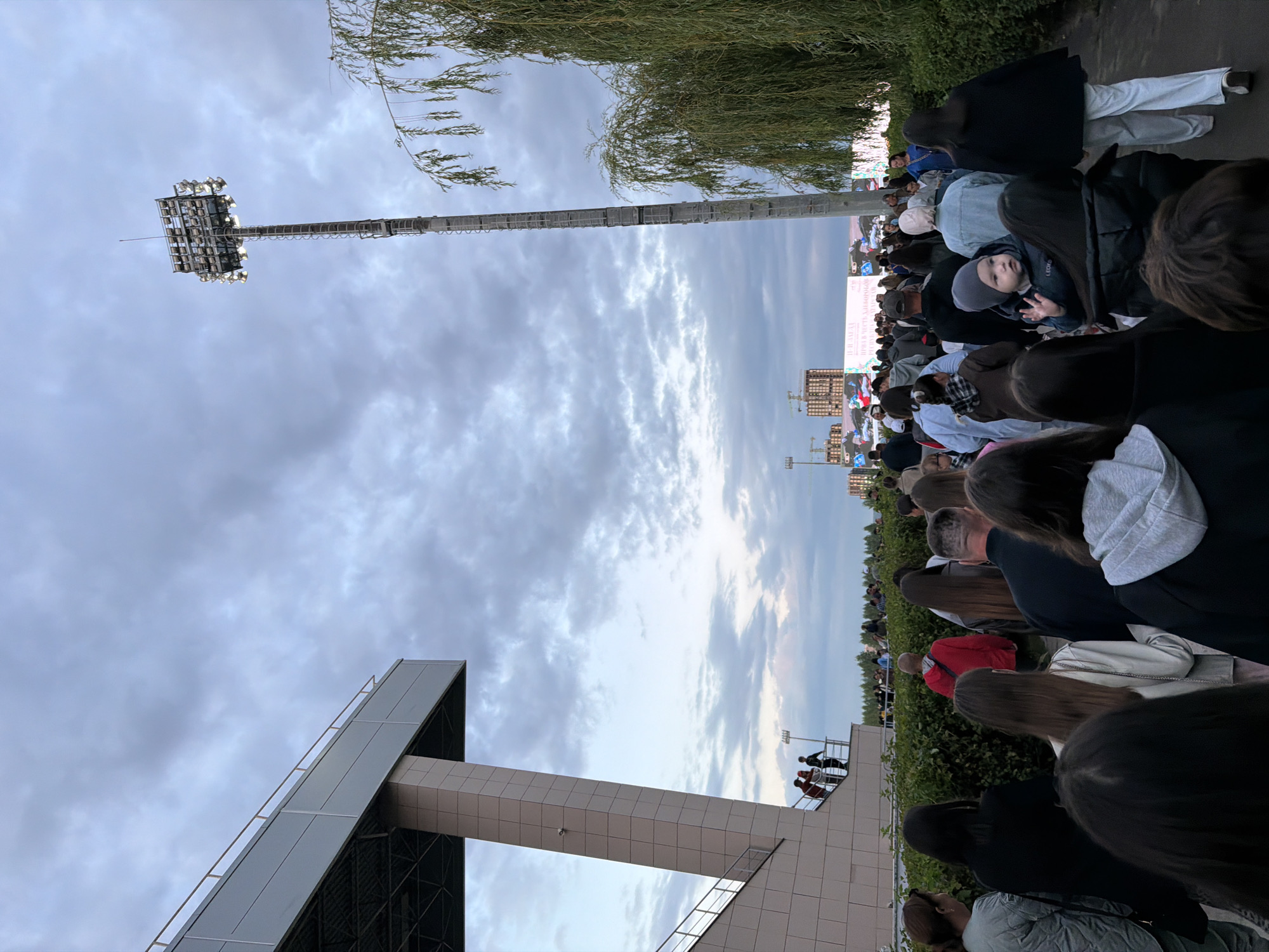 The image depicts a large gathering of people in an outdoor urban setting, possibly a stadium or event venue, under a cloudy sky. In the foreground, a crowd of individuals are walking in the same direction. The people are diverse in age and attire, with some individuals carrying bags or wearing jackets. Behind the crowd, a large stadium structure with a slanted roof and supporting pillars is visible on the left. On the right side of the image, a tall floodlight tower illuminates the scene, casting light on the surrounding trees and vegetation. In the distance, city buildings and construction cranes can be seen, indicating an urban environment. A large screen or display is also visible in the background, suggesting a planned event. The overall atmosphere appears to be one of anticipation or movement towards an event.