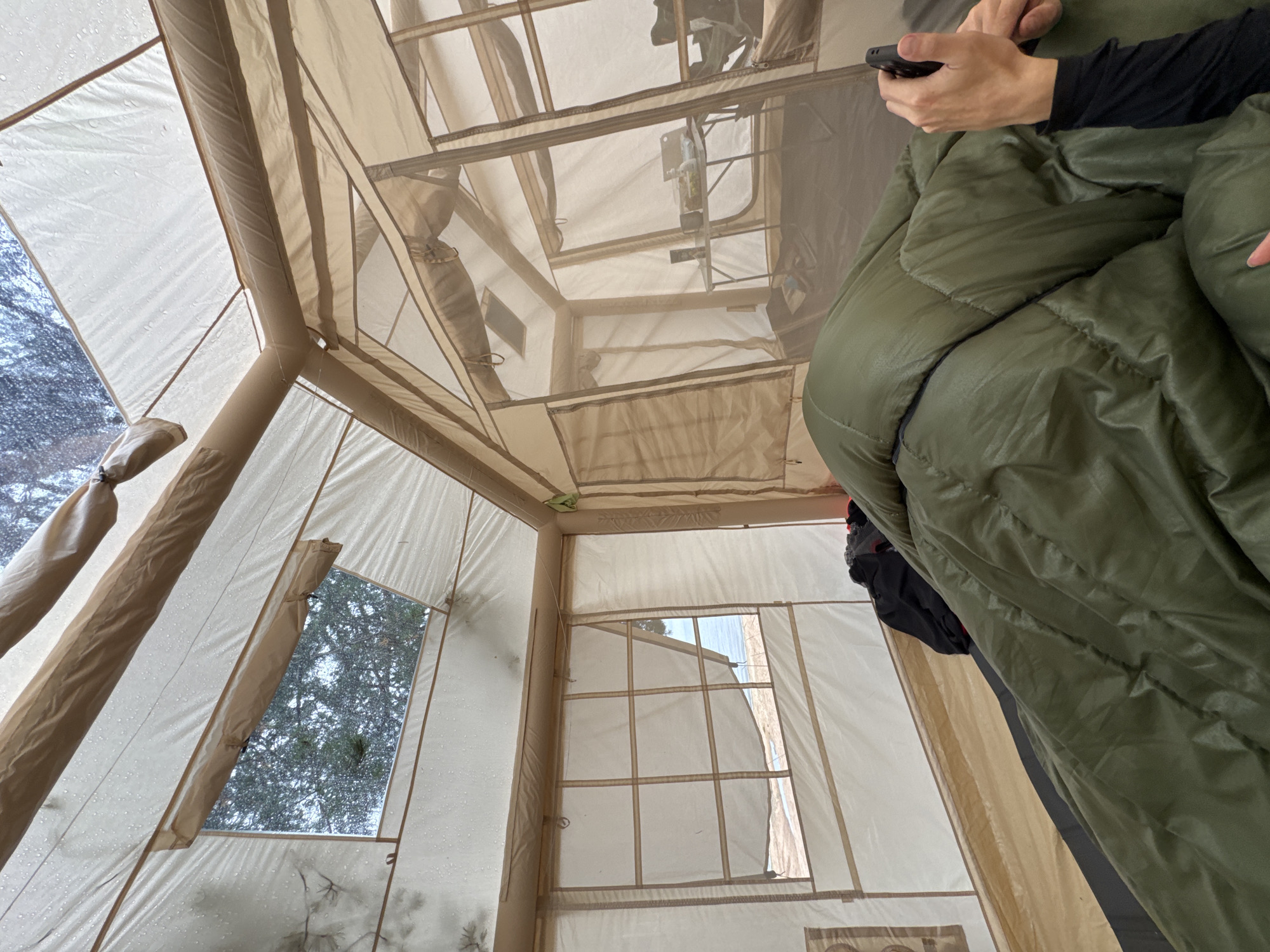 The image shows the interior of a large canvas tent with multiple rooms and screened windows. A person is lying in a green sleeping bag in the foreground, holding a dark-colored phone in their right hand. The person is wearing a black long-sleeved shirt. In the background, there is a small table with items on it, including containers and what appears to be a bottle. The tent walls are tan, and the ceiling is made of a lighter fabric. Outside the screened windows, trees and water are visible, suggesting a natural setting. The tent appears to be set up in a campsite.