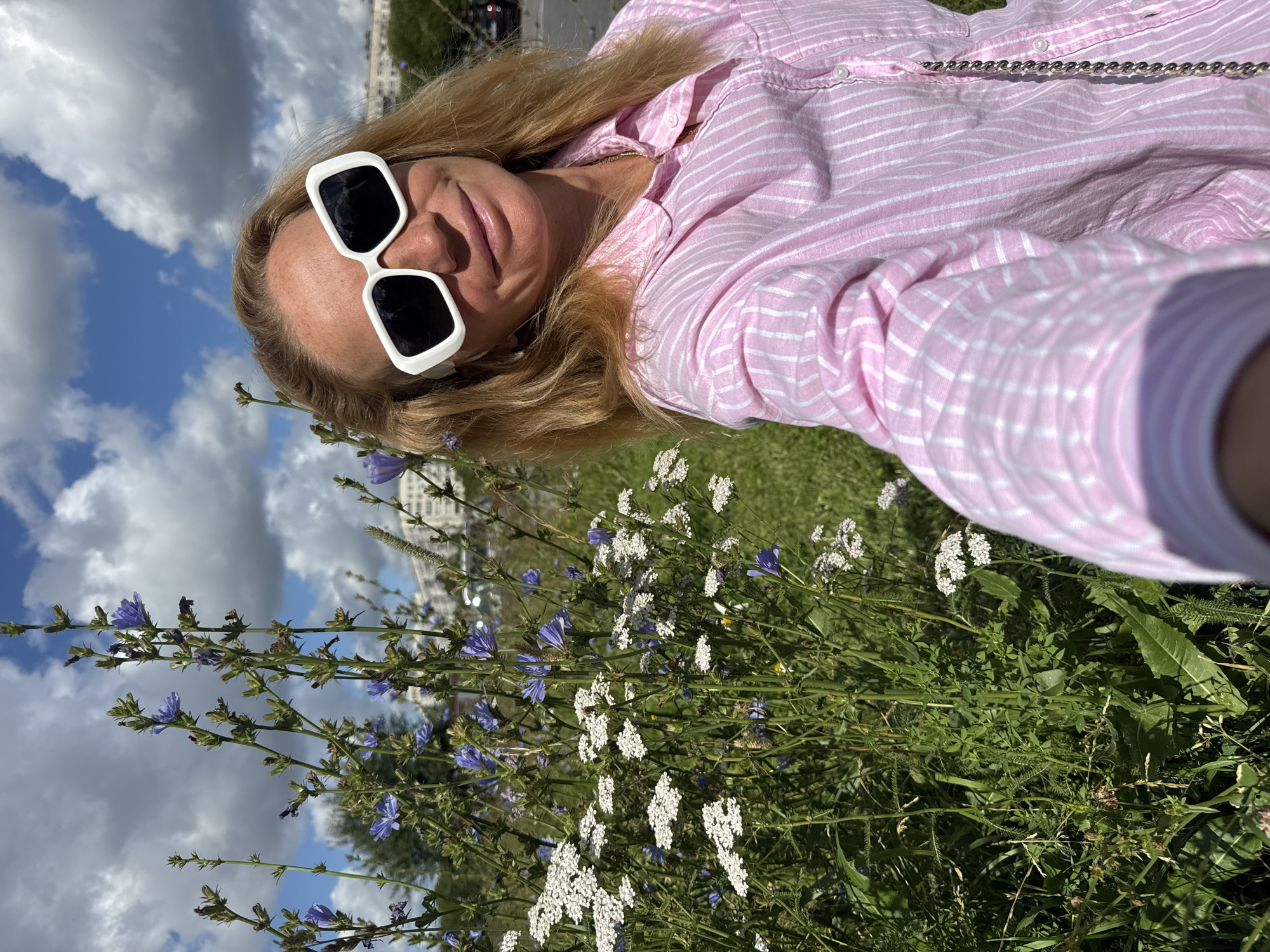 A woman wearing white-framed sunglasses and a pink and white striped button-down shirt is taking a selfie outdoors. She is smiling and looking at the camera. In the background, there are tall green plants with small purple and white flowers, as well as a partly cloudy blue sky. A distant cityscape with buildings is visible on the right side of the image. The woman appears to be in an urban park or green space in Saint Petersburg, Russia, given the characteristic architecture and vegetation.