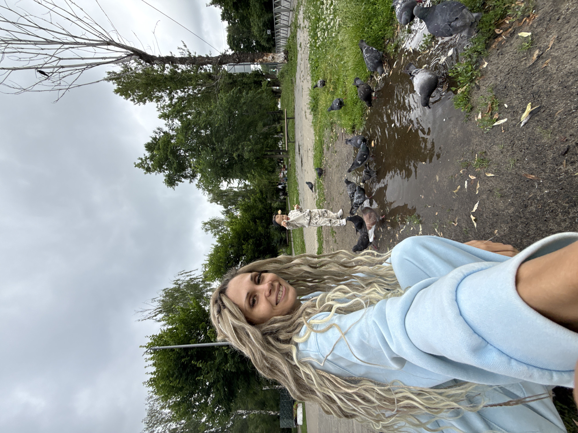 A low-angle, wide shot shows a woman with long, blonde dreadlocks smiling at the camera. She is wearing a light blue tracksuit and is kneeling on the ground. In the background, a young child in a light-colored outfit stands in front of a puddle, surrounded by pigeons. The child appears to be feeding the birds. The scene is set in a park-like environment with green trees and grass, and a cloudy sky overhead. A dead tree with a bird perched on it is visible to the right.