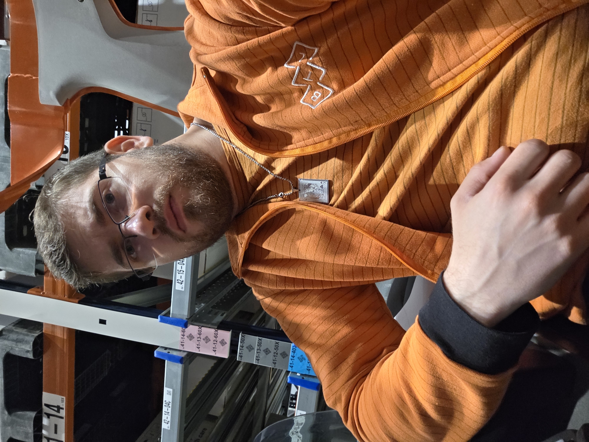 A man with a beard and glasses, wearing an orange striped zip-up jacket, stands in front of metal shelving racks. He has a silver chain around his neck with a rectangular pendant. The racks have labels with codes such as "42-15-04" and "41-14-60X". The codes seem to denote shelf or product IDs. The image contains no specific clues indicating the location is in Plovdiv, Bulgaria.