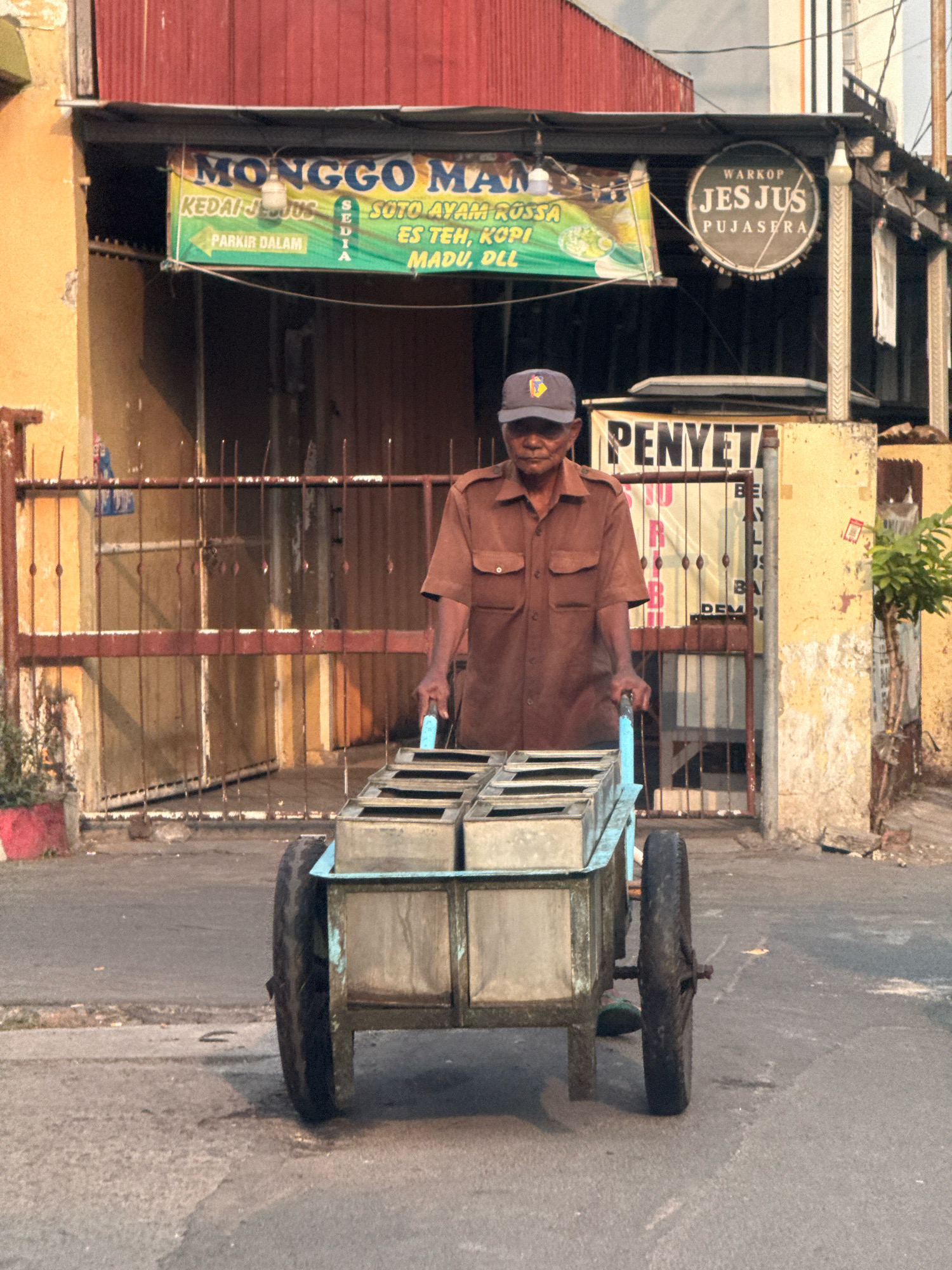 An elderly Indonesian man with weathered skin and wearing a brown uniform and a dark cap pushes a cart filled with aluminum containers down a street in Surabaya, Indonesia. The cart has two large wheels and a blue frame. Behind the man, a red corrugated metal building with a black awning is visible. A green banner hangs from the awning with Indonesian text that includes "MONGGO MAN", "KEDAI", "SOTO AYAM", "ES TEH", and "PARKIR DALAM". To the right of the banner, a circular sign reads "WARKOP JES JUS PUJASERA". Another banner with Indonesian text is partially visible behind the man, reading "PENYET". The street is paved with asphalt, and there is a sidewalk with some vegetation on the right side of the image. The scene appears to be captured during daylight, with the sun casting shadows on the ground.