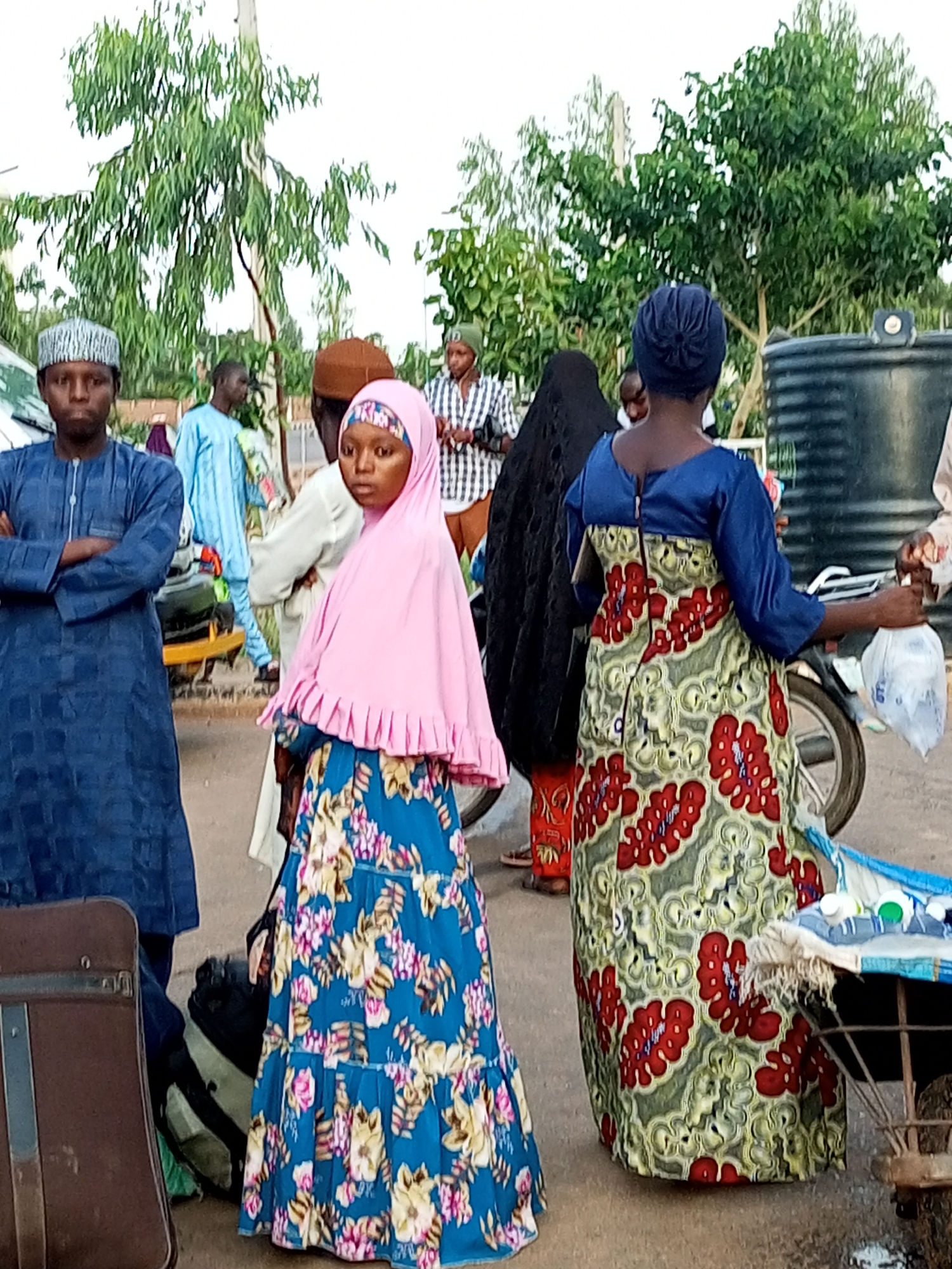 In the city of Gombe, Nigeria, a group of people are gathered near a street. On the left, a man wearing a blue traditional outfit and a patterned cap stands with his arms crossed. To his right, a young girl in a pink hijab and a floral blue dress is looking to the side. Behind her, a woman in a blue headwrap and a green and red patterned dress is facing away from the camera, holding a plastic bag. Other individuals are visible in the background, some wearing traditional attire. The environment appears to be an outdoor setting with trees and a large black water tank visible. Luggage, including a brown suitcase, is present on the ground, suggesting travel. A cart with some items on it is also in the foreground. The lighting indicates it may be daytime.