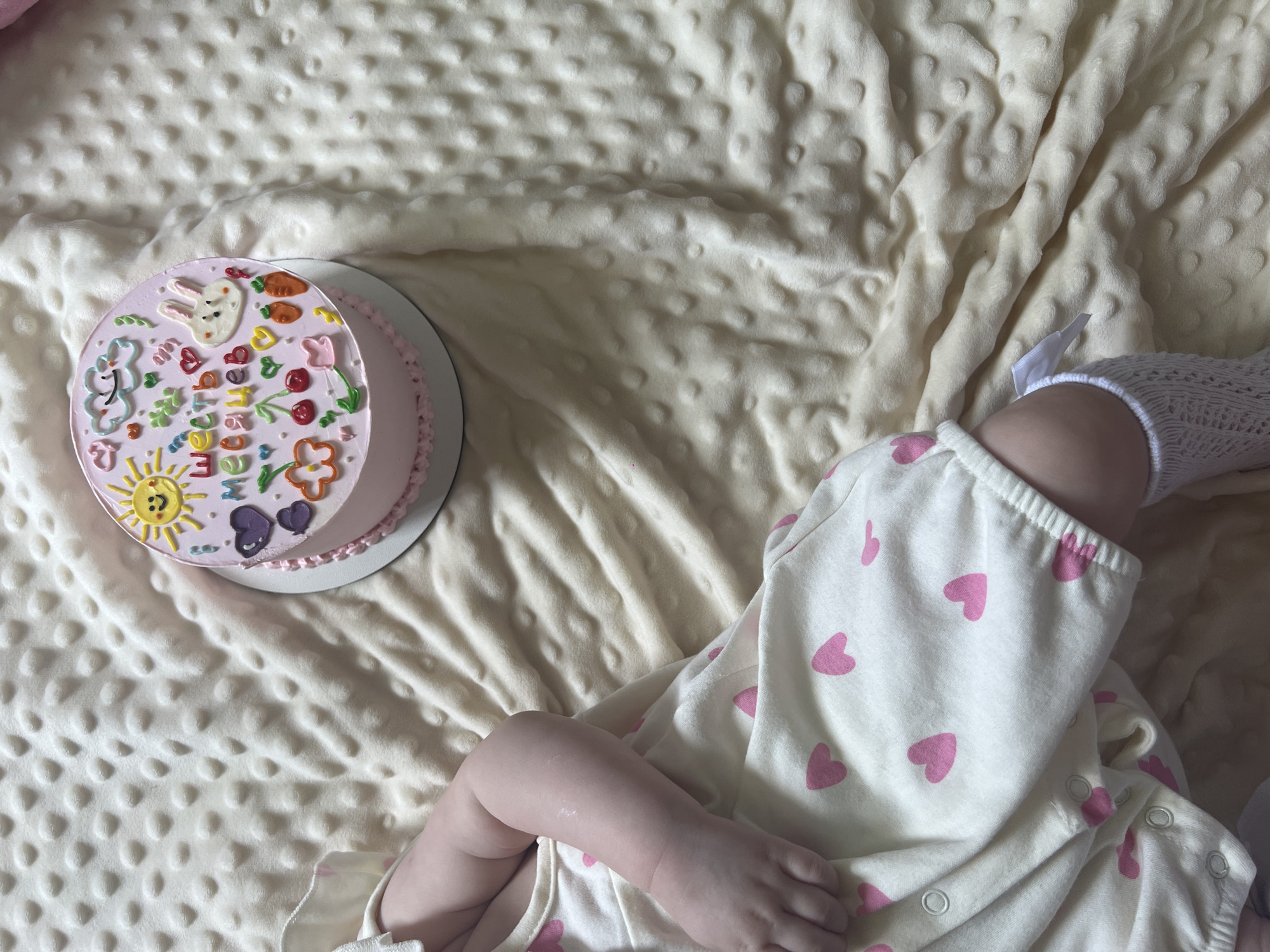 A baby lies on a light yellow blanket with a cake next to it. The baby is wearing a white onesie with pink hearts and white crocheted socks. The cake is round with pink frosting and has decorations of a bunny, carrots, flowers, and the word "HOME". The scene is lit by natural light. The location is Chaikovsky, Russia.