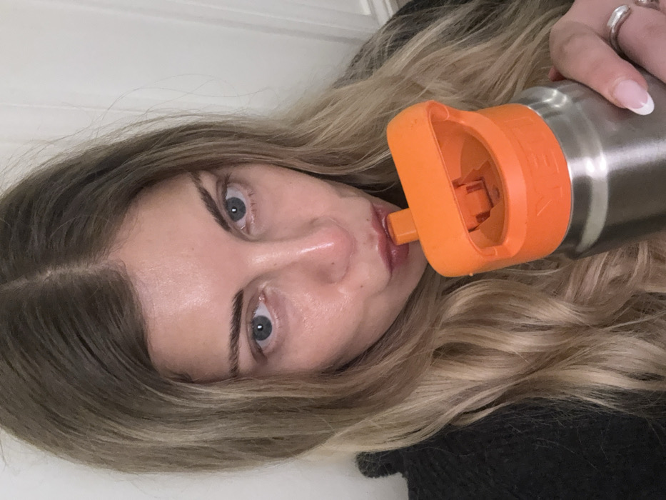 A blonde woman with fair skin drinks from a silver water bottle with an orange lid. Her eyes are blue, and she has manicured nails. She is facing the camera at an angle, and the background is a white wall. She is wearing a black shirt.