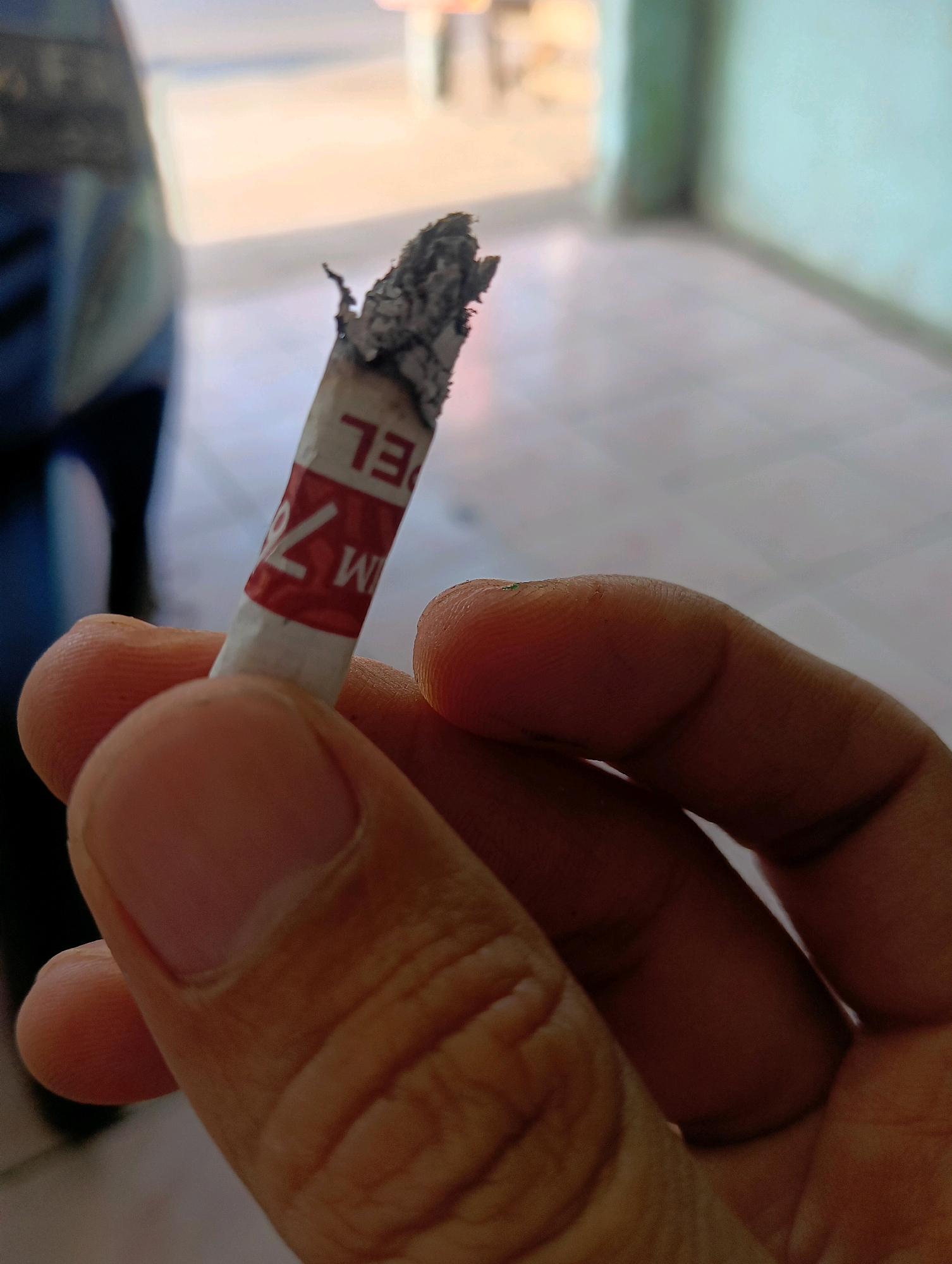 A close-up, eye-level shot captures a hand holding a partially smoked cigarette. The cigarette, positioned vertically in the frame, features a white filter and a red band with white text that is partially visible and upside down, reading "EL" and "IM 7%". The tip of the cigarette is ashed. The hand, with visible fingerprints and skin texture, is holding the cigarette between the thumb and index finger. The background is softly blurred, suggesting an indoor or semi-outdoor setting with muted colors. A dark, out-of-focus object is visible to the left, and a light-colored wall with a green tint occupies the right side of the background. The lighting is soft, casting subtle shadows. This image was taken in Sokaraja Kidul, Indonesia.