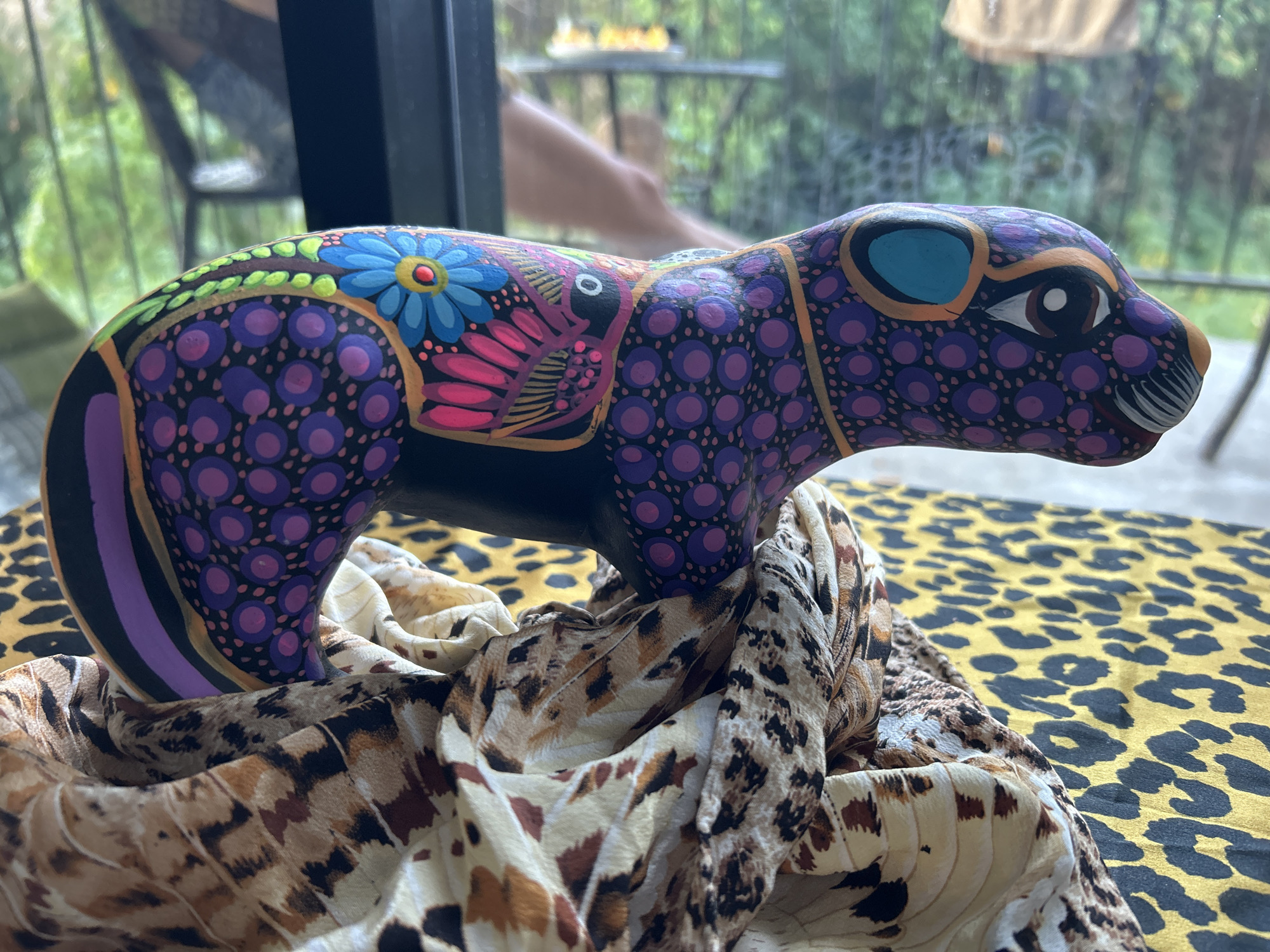 The image shows a brightly painted wooden sculpture of a cat-like animal, possibly a jaguar or ocelot, resting on a patterned scarf. The sculpture is painted with vibrant colors and intricate designs, including floral motifs and abstract patterns of purple dots. The scarf appears to be a mix of animal print patterns, possibly leopard and snake. The background is blurry but suggests an outdoor setting with greenery visible through a window or doorway. There is a railing visible and the legs of a person in the background. The location is not explicitly stated in the image or the context.