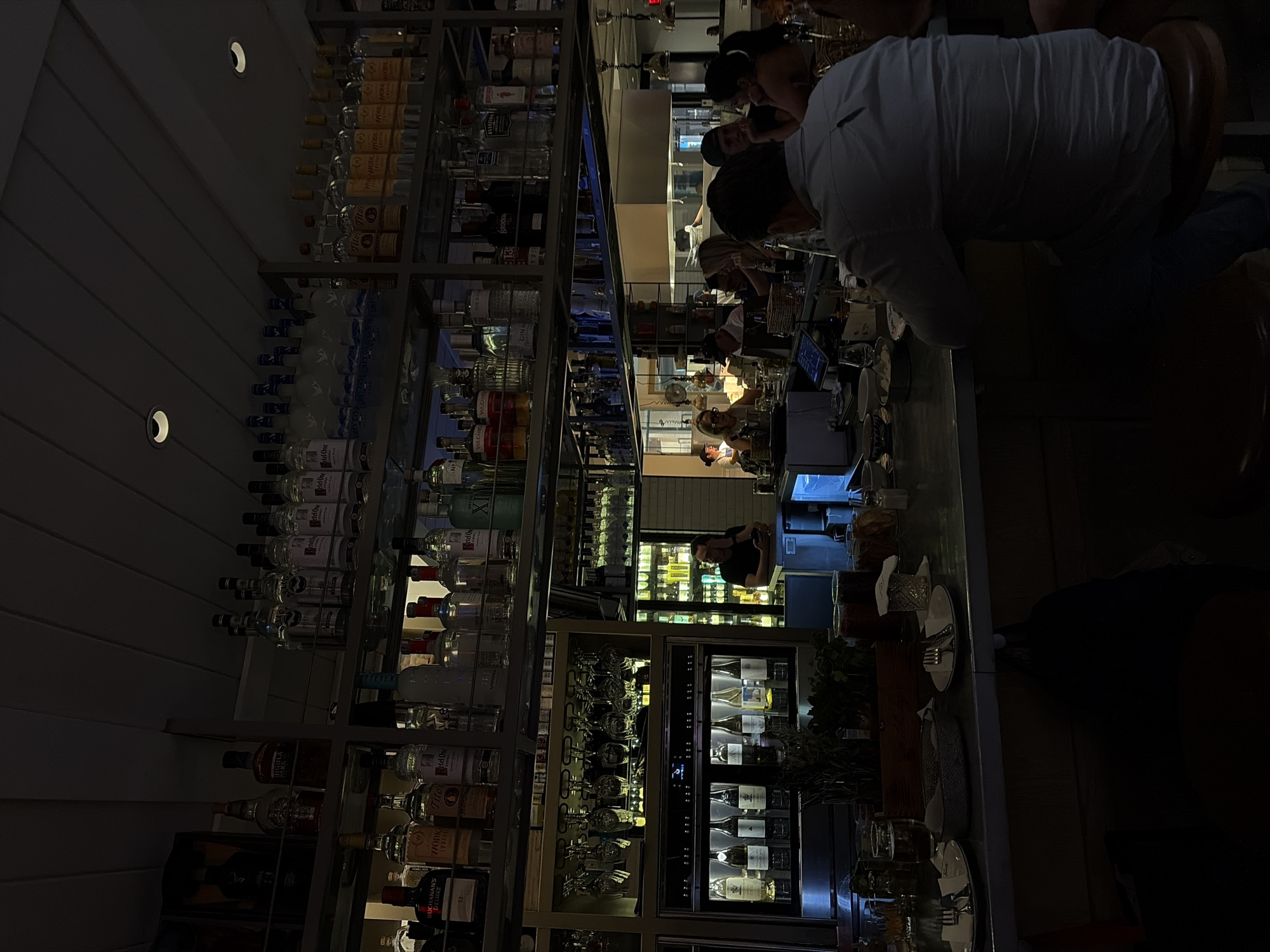 The scene depicts the dimly lit interior of a bar, likely located in Miami, United States of America, given the style of establishment and ambiance common to the city. Patrons are seated at the bar counter, and a bartender is visible in the background, presumably preparing drinks. The bar's back wall features an extensive display of liquor bottles arranged on shelving units, indicating a well-stocked establishment. In addition, a separate unit displays wine bottles and glassware. The overall atmosphere is one of a relaxed, social setting with patrons enjoying their time at the bar. A partially visible dark object is located close to the lower-right edge.
