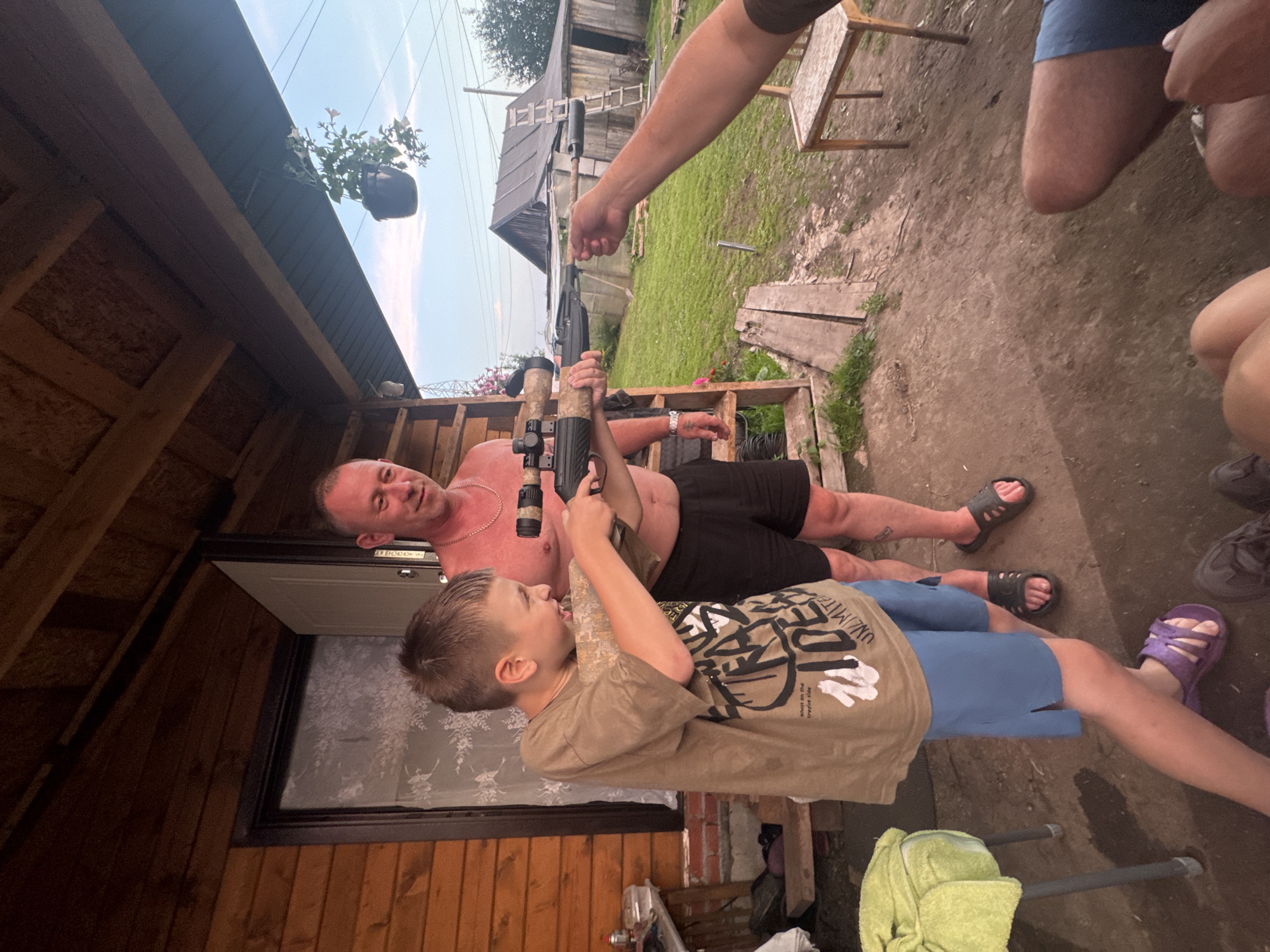 In Chaikovsky, Russia, a young boy, wearing a tan t-shirt and blue shorts, aims a rifle with a scope. A shirtless man in black shorts stands behind him, smiling. Another person's arm extends into the frame, holding the rifle's barrel. They are on a wooden porch of a log cabin, with a grassy yard and a simple wooden building in the background. The scene appears to be in a rural or suburban setting.