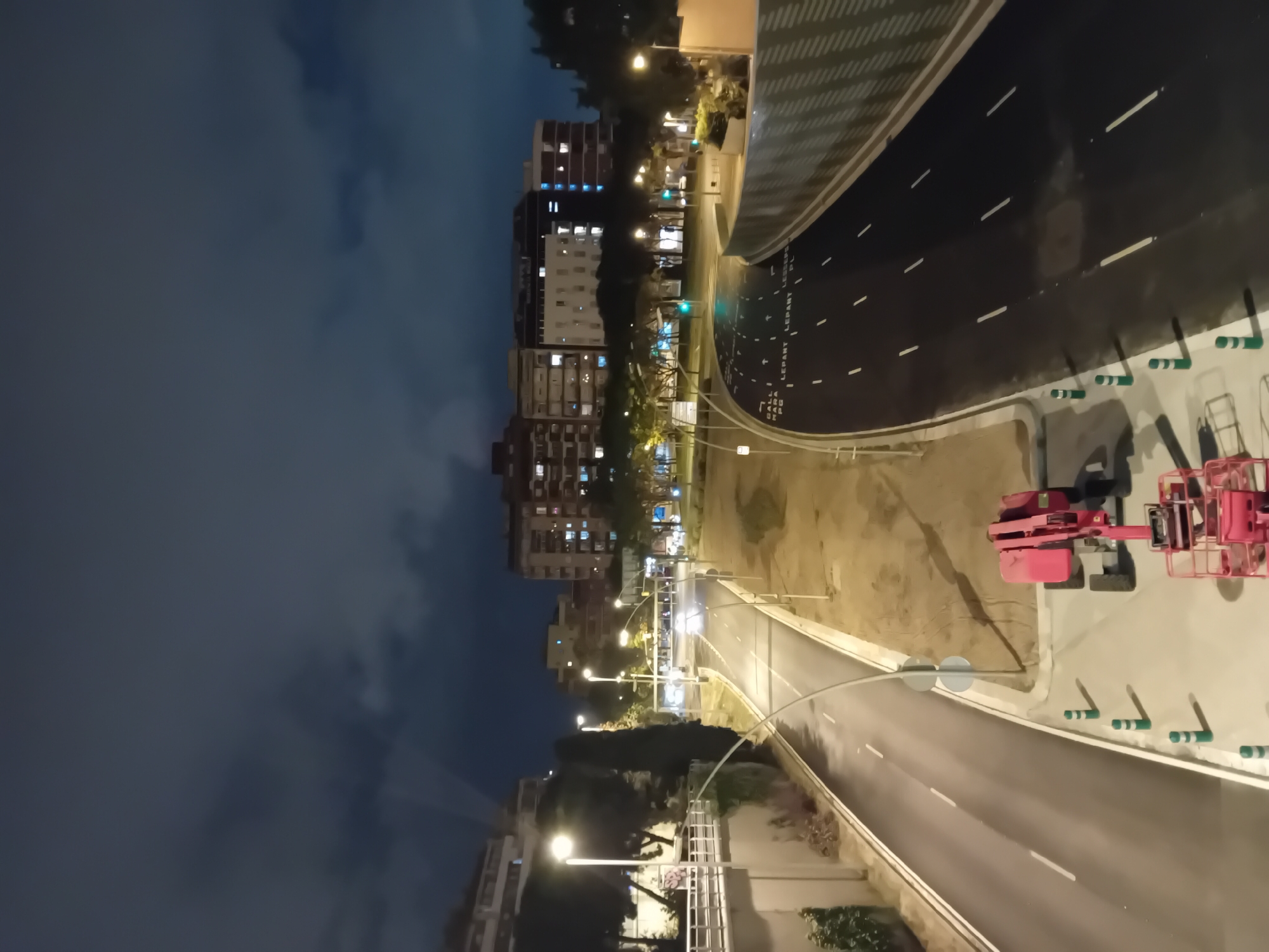 The image shows a nighttime view of a street in Barcelona, Spain, identified by the street names "HALL" and "LEPANT" painted on the pavement. A newly paved or resurfaced roadway curves to the right, featuring fresh white lane markings. Near the upper right corner are two pieces of pink construction equipment parked on a paved surface. A concrete barrier with green and white striped markers runs along the edge of the roadway. Streetlights illuminate the scene, and buildings with lit windows are visible in the background on the left side of the image. The sky above is dark, indicating it is nighttime. The photograph appears to be taken from an elevated position, possibly a building or bridge.