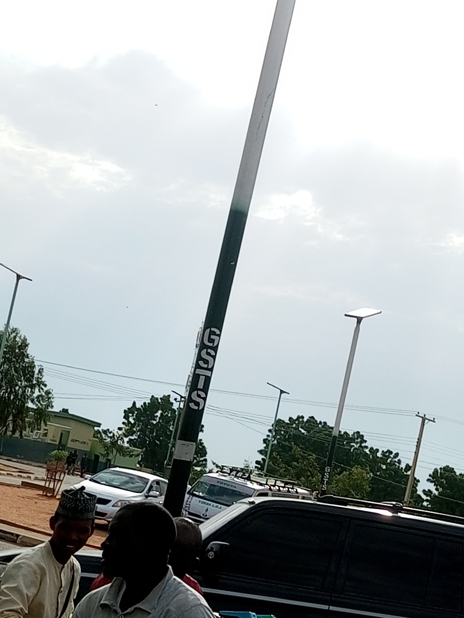 The image captures a street scene in Gombe, Nigeria, under an overcast sky, suggesting it might be late morning or early afternoon. The foreground is dominated by the dark silhouette of a large vehicle, likely an SUV, with its roof rack visible. In front of this vehicle, two men are the primary human subjects. One man, on the left, is wearing a light-colored traditional outfit and a patterned cap, and he is looking towards the camera with a slight smile. The other man, on the right, is mostly in silhouette, with his head and shoulders visible, wearing a collared shirt.
Behind the vehicles, several trees with lush green foliage line the street. In the mid-ground, a white sedan is parked. Further back, a building with visible windows and an entrance is present, giving a sense of an urban or semi-urban environment. Several streetlights are visible throughout the scene, including a prominent, tall pole on the right side of the image with the letters "GSIS" vertically inscribed on it in white. Another streetlight with a distinctive modern design is visible on the left side of the image. Power lines stretch across the background, typical of many Nigerian streetscapes. The overall impression is of a casual gathering or a moment paused on a busy street.