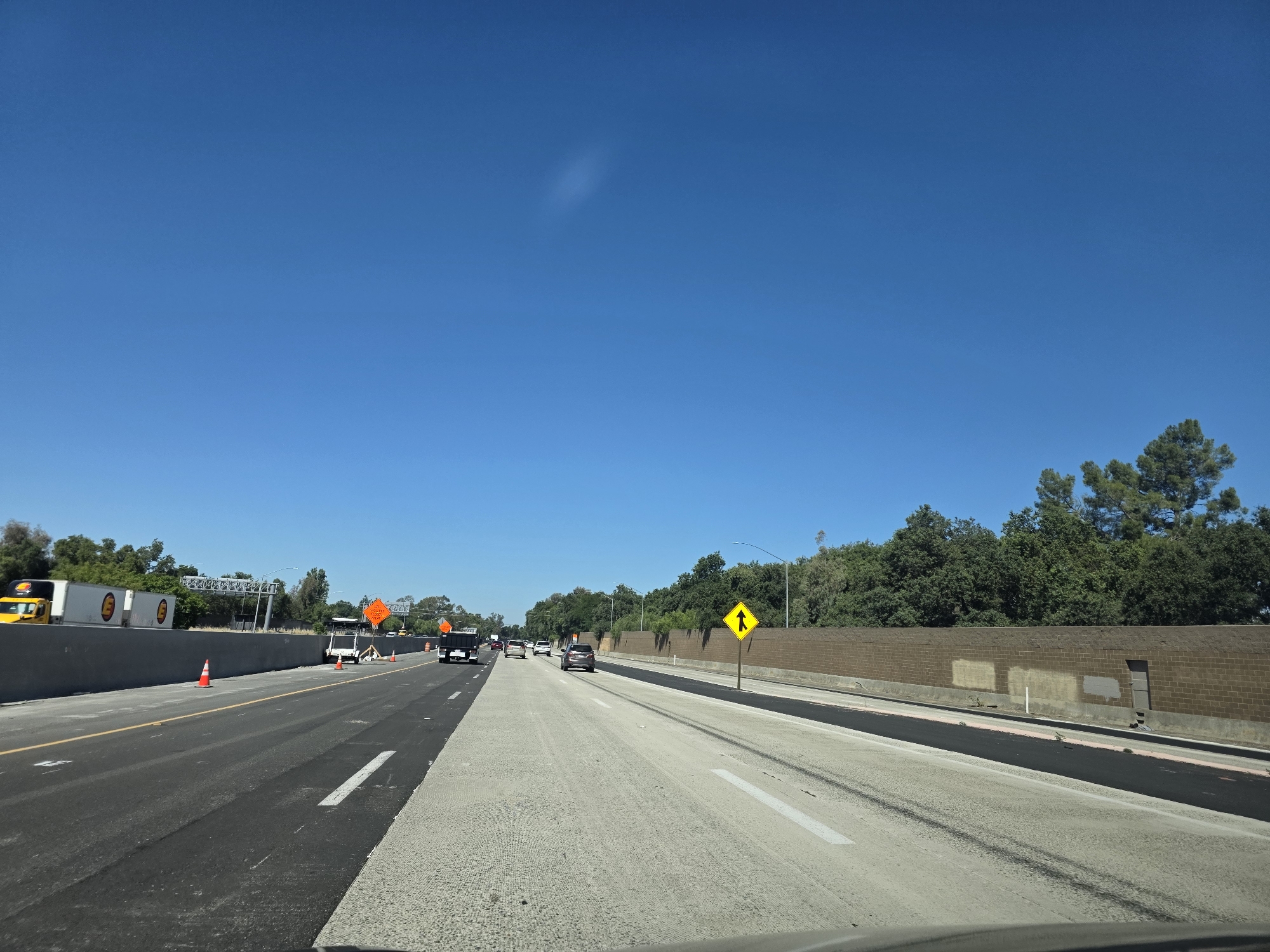 The image captures a view of a highway with ongoing construction in West Sacramento, California. The highway is divided into multiple lanes, with some sections appearing freshly paved with asphalt and others with a concrete surface. Traffic cones are visible along the sides of the road, indicating lane closures or construction zones. Several vehicles, including cars and trucks, are on the highway, navigating the construction. On the right side of the highway, a brick wall serves as a barrier, with a door set into it. Beyond the wall, lush green trees form a dense line, suggesting a natural landscape bordering the urban area. The sky above is a clear, vibrant blue, indicating a sunny day. The overall scene depicts a typical highway environment with the presence of construction and road work.