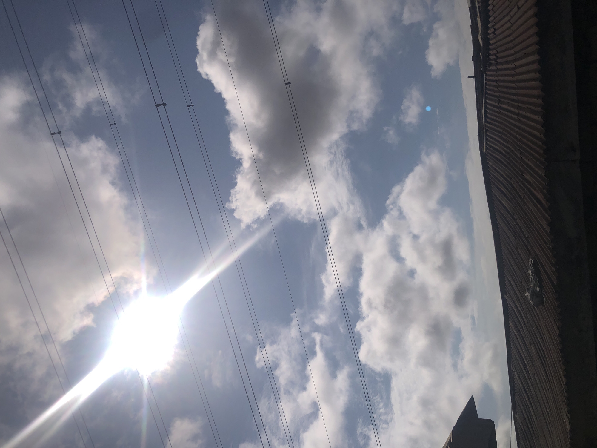 A low-angle, vertical shot captures a bright sun with distinct rays angled downwards and to the left, partially obscured by clouds. Multiple horizontal power lines stretch across the frame, with insulators visible on some. Below the power lines and clouds, a corrugated metal roof forms the lower third of the image, with a dark, indistinct structure to the far left. The scene appears to be taken outdoors in the city of Ajah, Nigeria, judging by the architecture and possibly the atmospheric conditions.