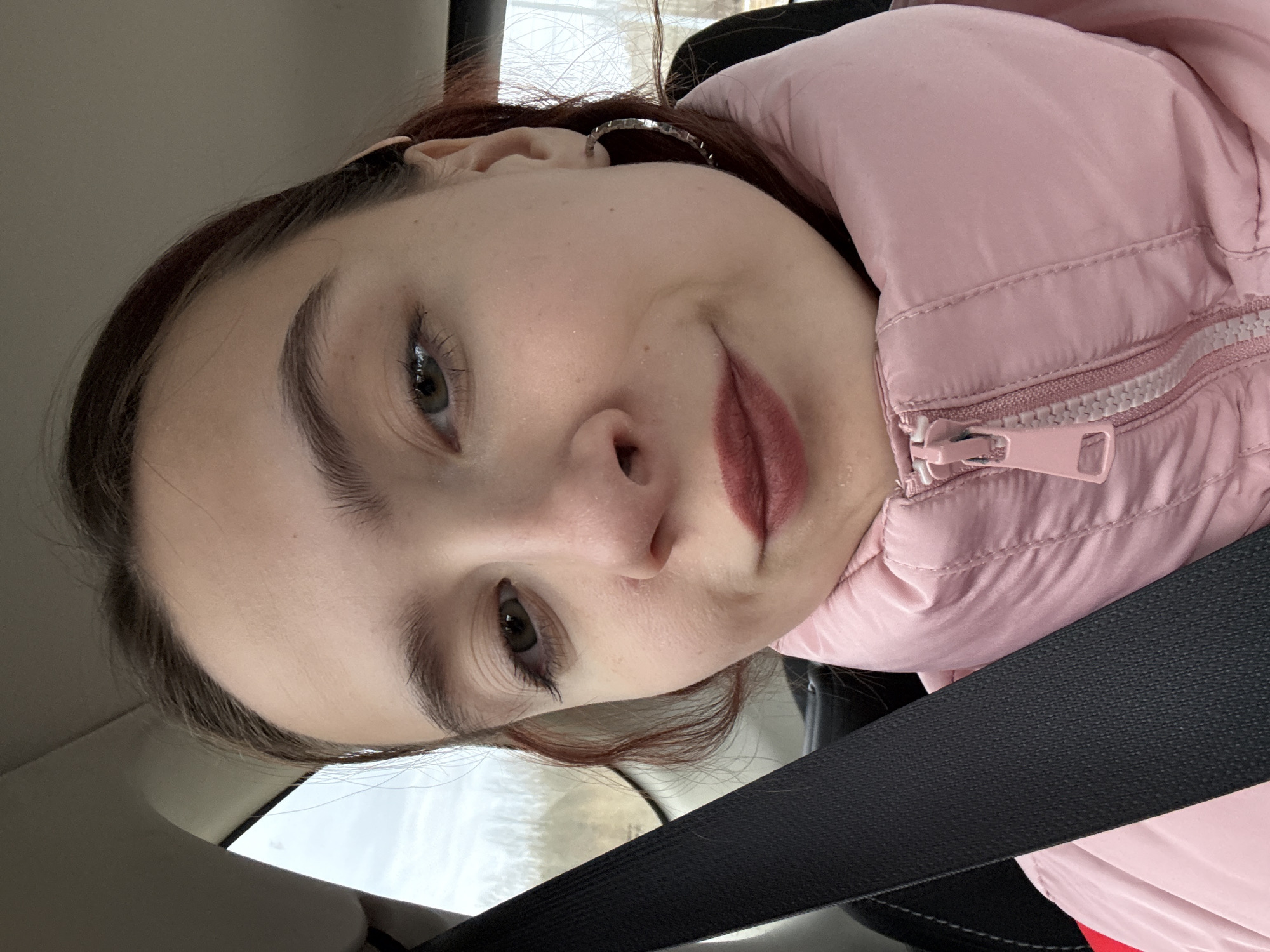 A person with brown hair and green eyes is in a car wearing a pink puffer jacket and a seatbelt. The background is blurry, showing snow and trees. The picture was not taken in Berezniki, Russia.